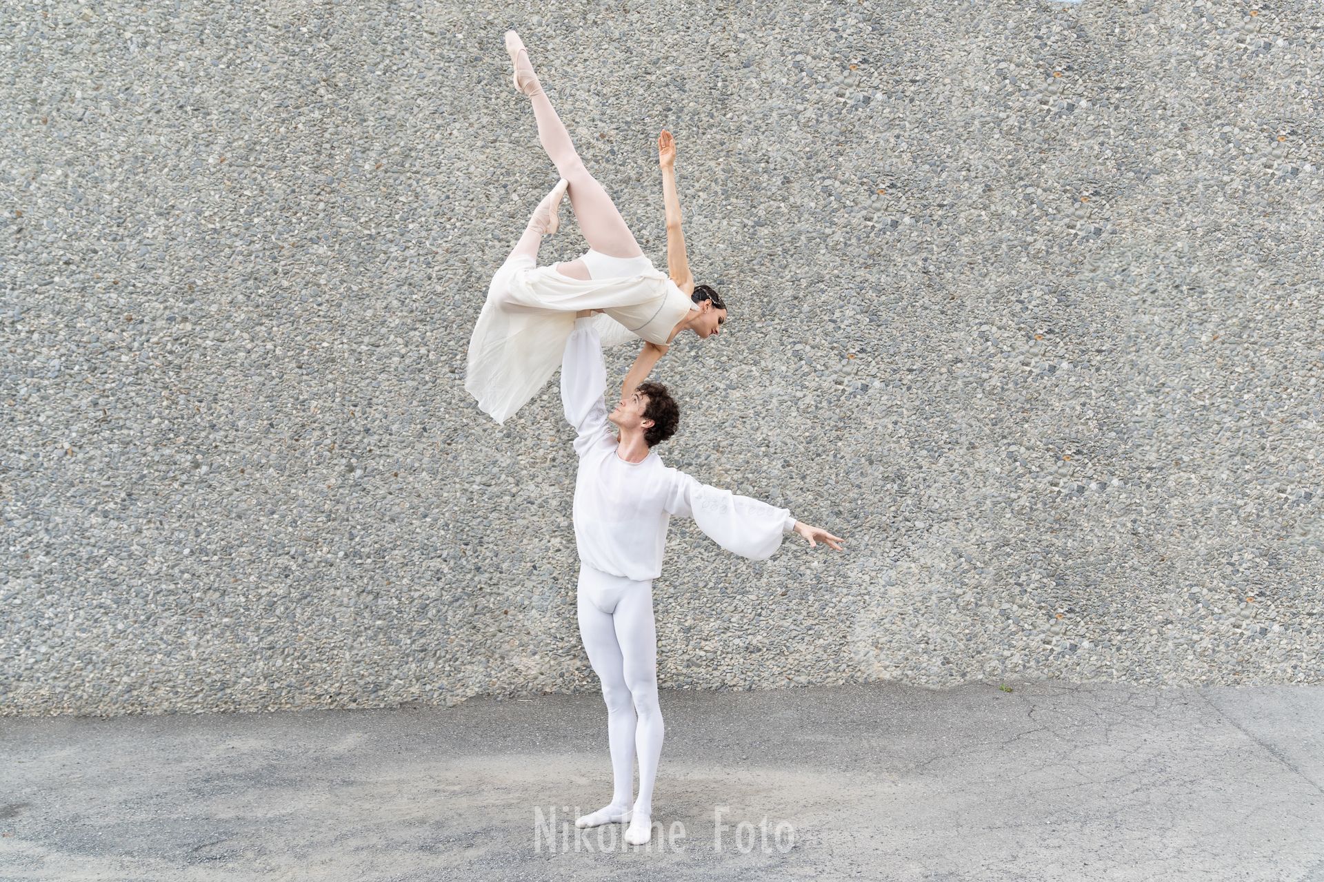 Ballet dancers in white attire perform a lift against a textured gray wall. The woman is in arabesque, arm overhead.