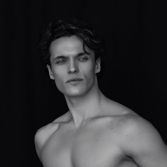 A shirtless man with wavy dark hair, looking off to the side, against a dark background.