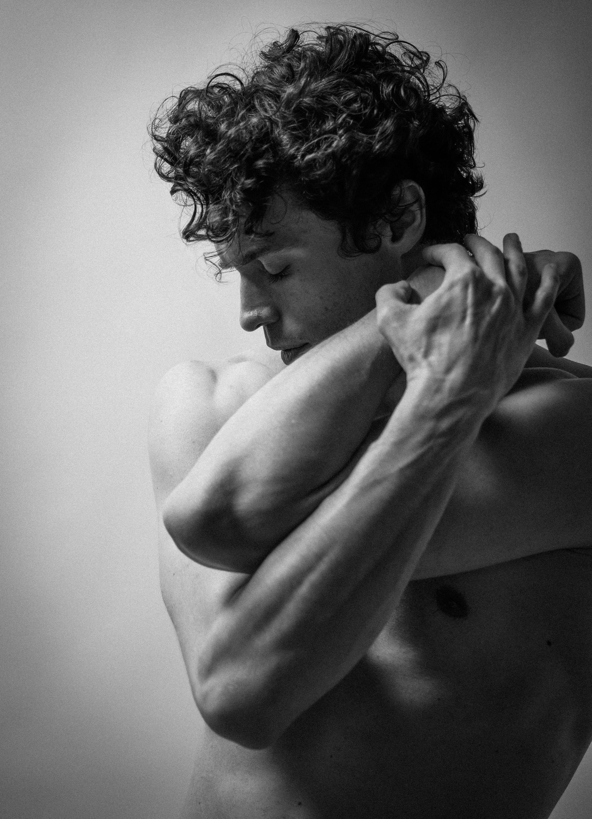 Man with curly hair stretches arms behind his neck, partially shirtless, in a studio setting.