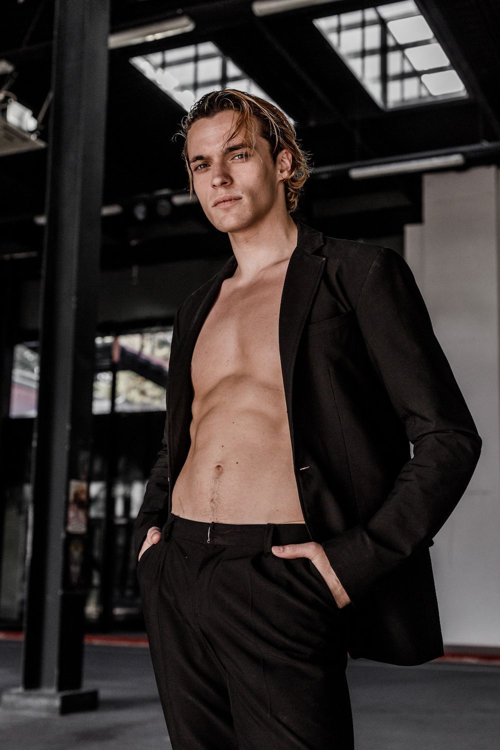 Man with abs in black suit, hand in pocket, standing in industrial setting.