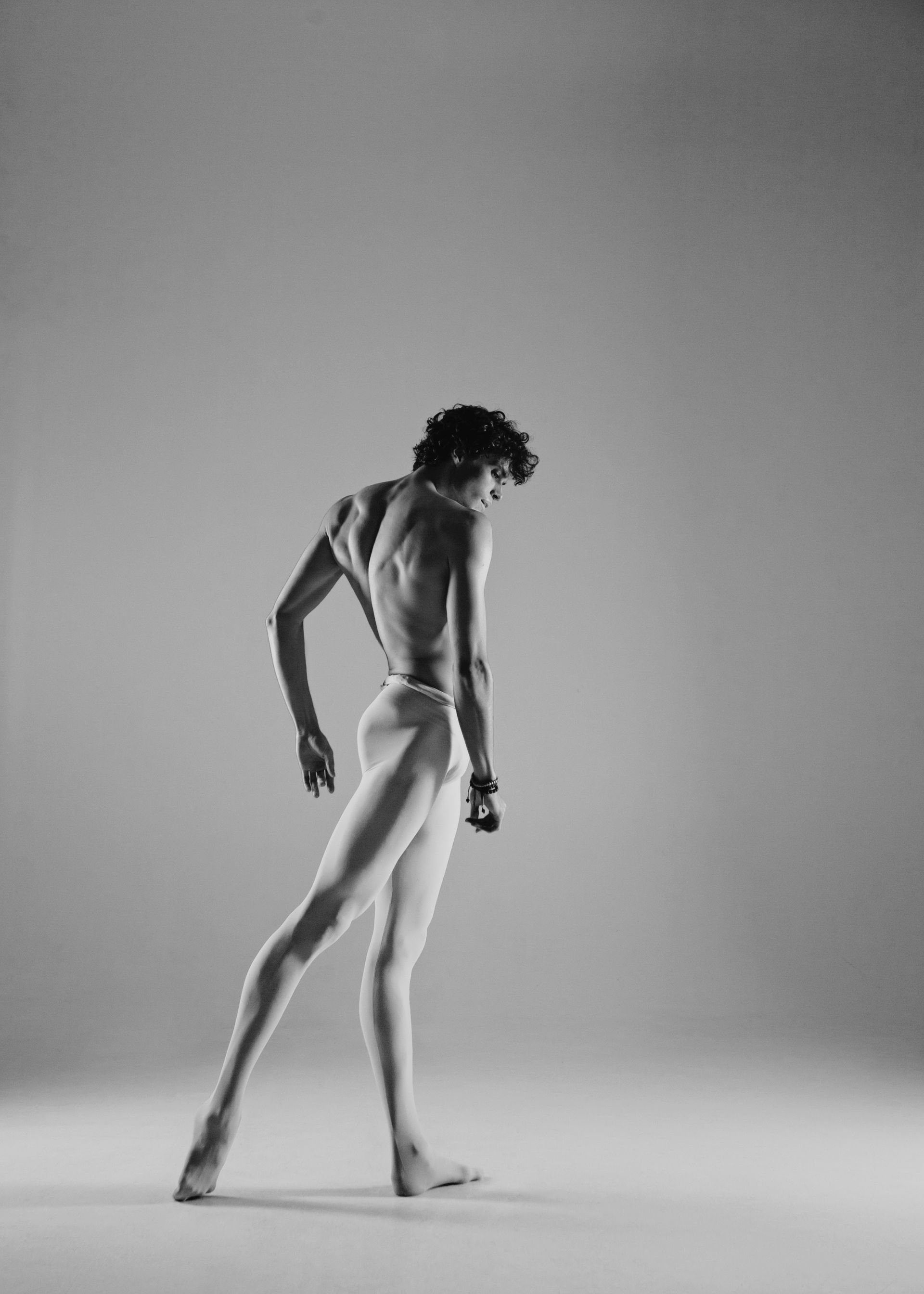 Ballet dancer in a white leotard, striking a pose with back arched, against a light gray background.