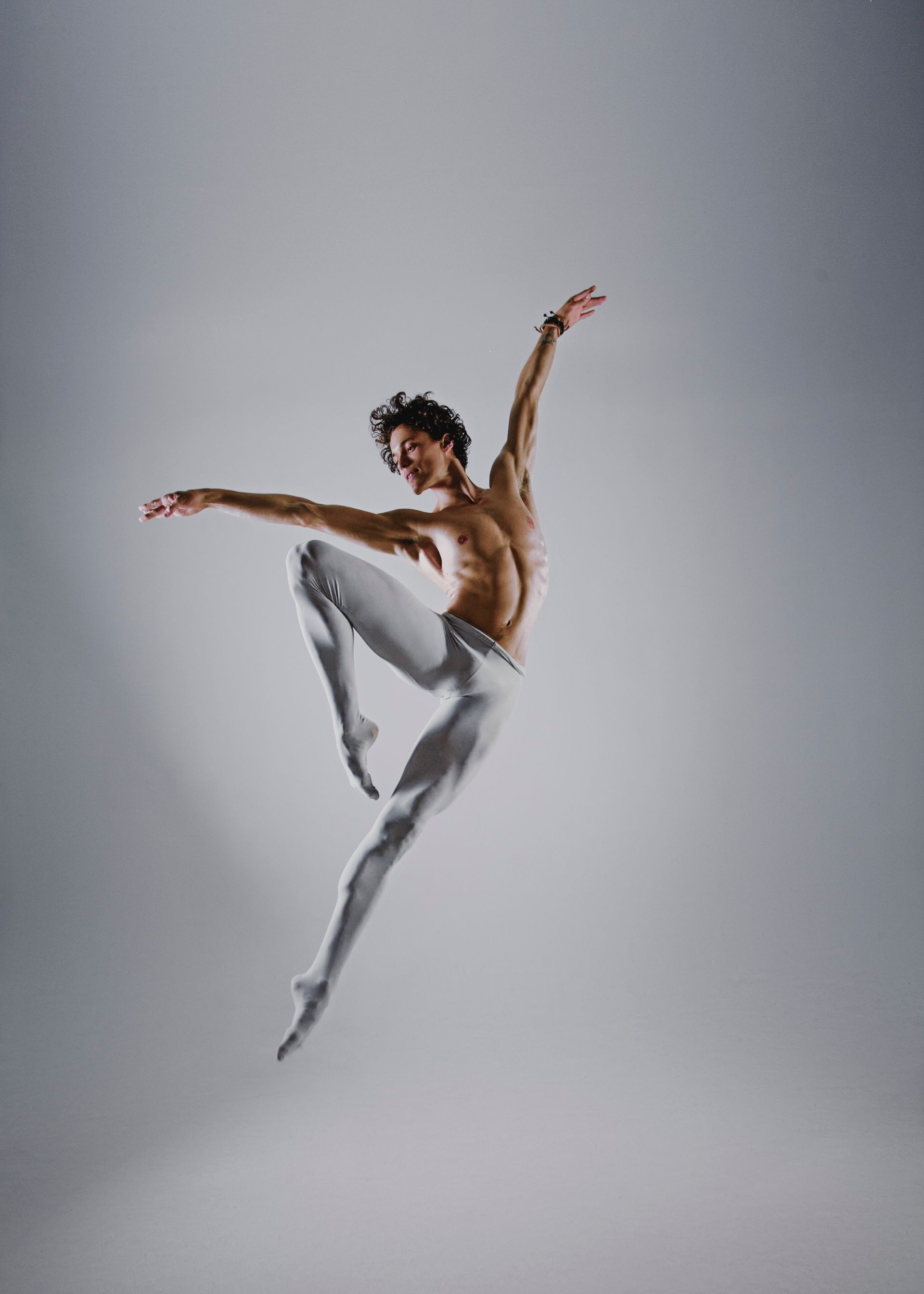 Ballet dancer leaps in the air, arms extended. Wearing white tights, he has a defined physique on a gray backdrop.