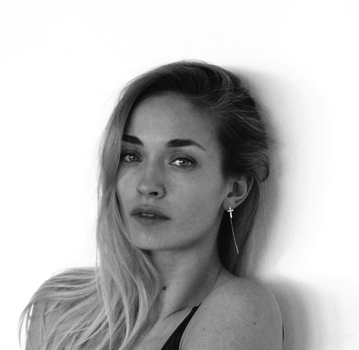 Woman leaning against a white wall, looking at the camera. Blonde hair, cross earring.