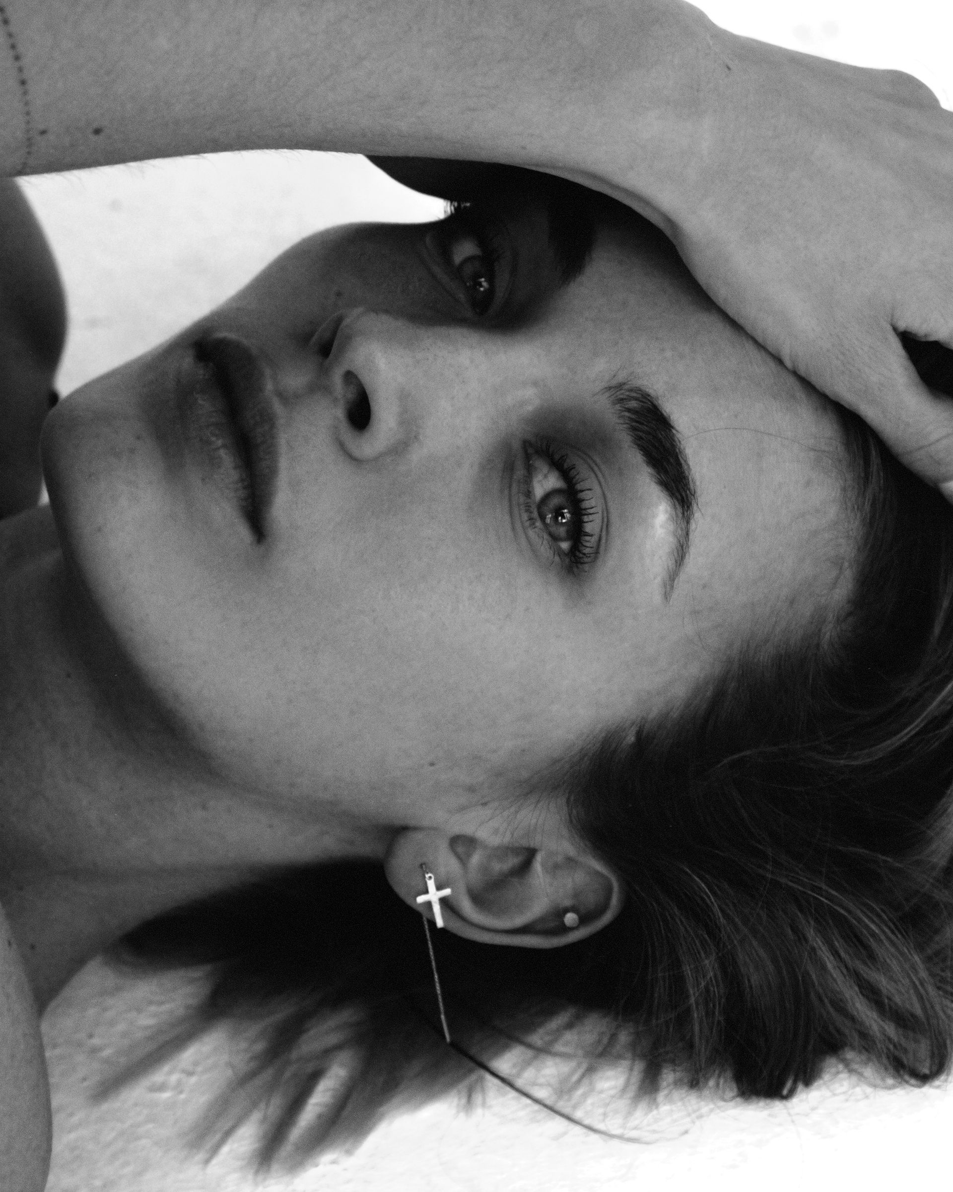 Woman with dark eye makeup, looking up, hand on forehead. Cross earring visible.