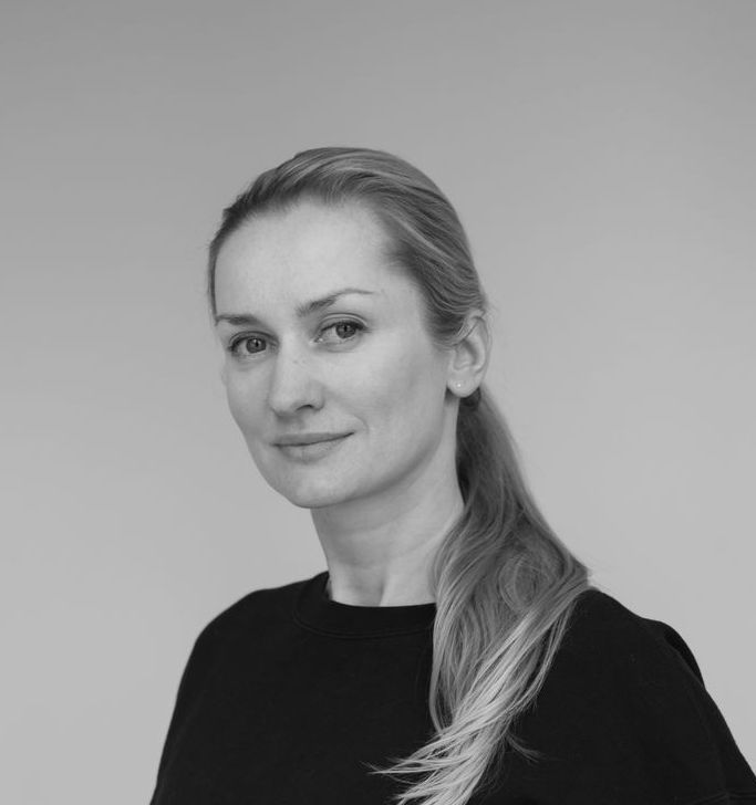 Woman with blonde hair pulled back in a low ponytail, smiling slightly, wearing a black top.