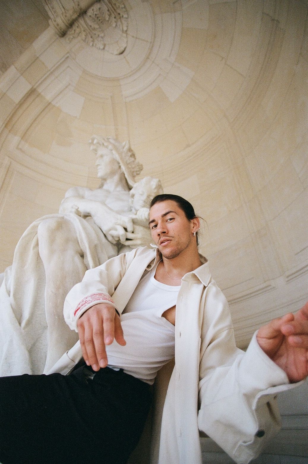 Man in white shirt and coat poses in front of a classical statue, reaching out with one hand.