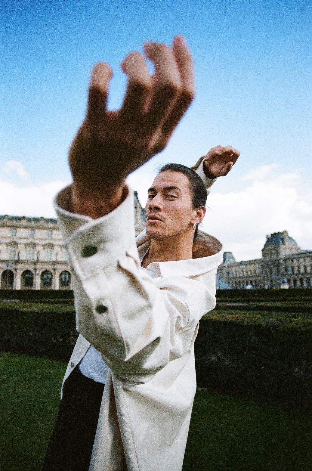 Person reaching up outdoors near the Louvre. Light-colored coat, blue sky, green grass.