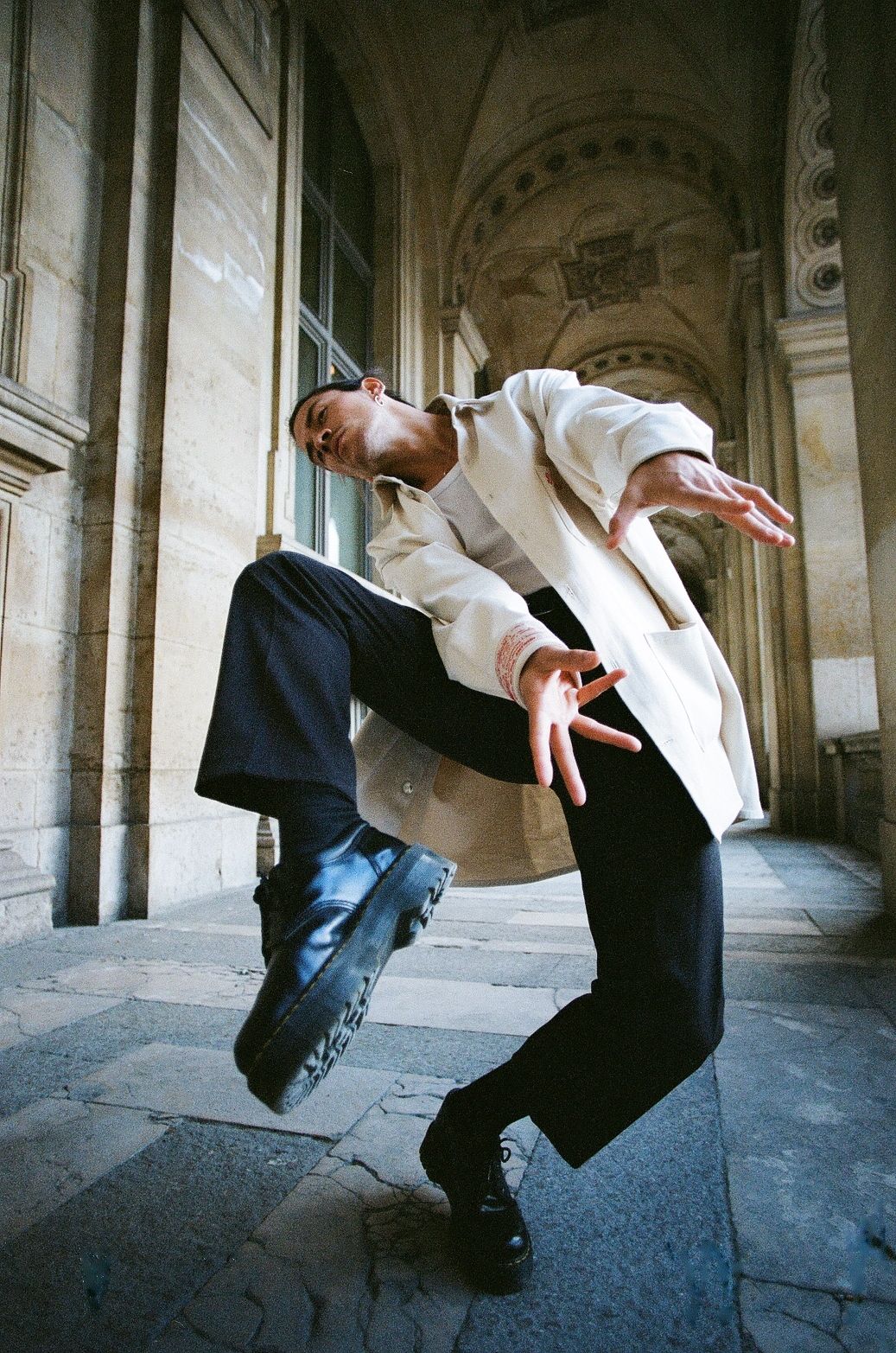Person in white jacket and black pants dances outdoors, raising arms and legs.