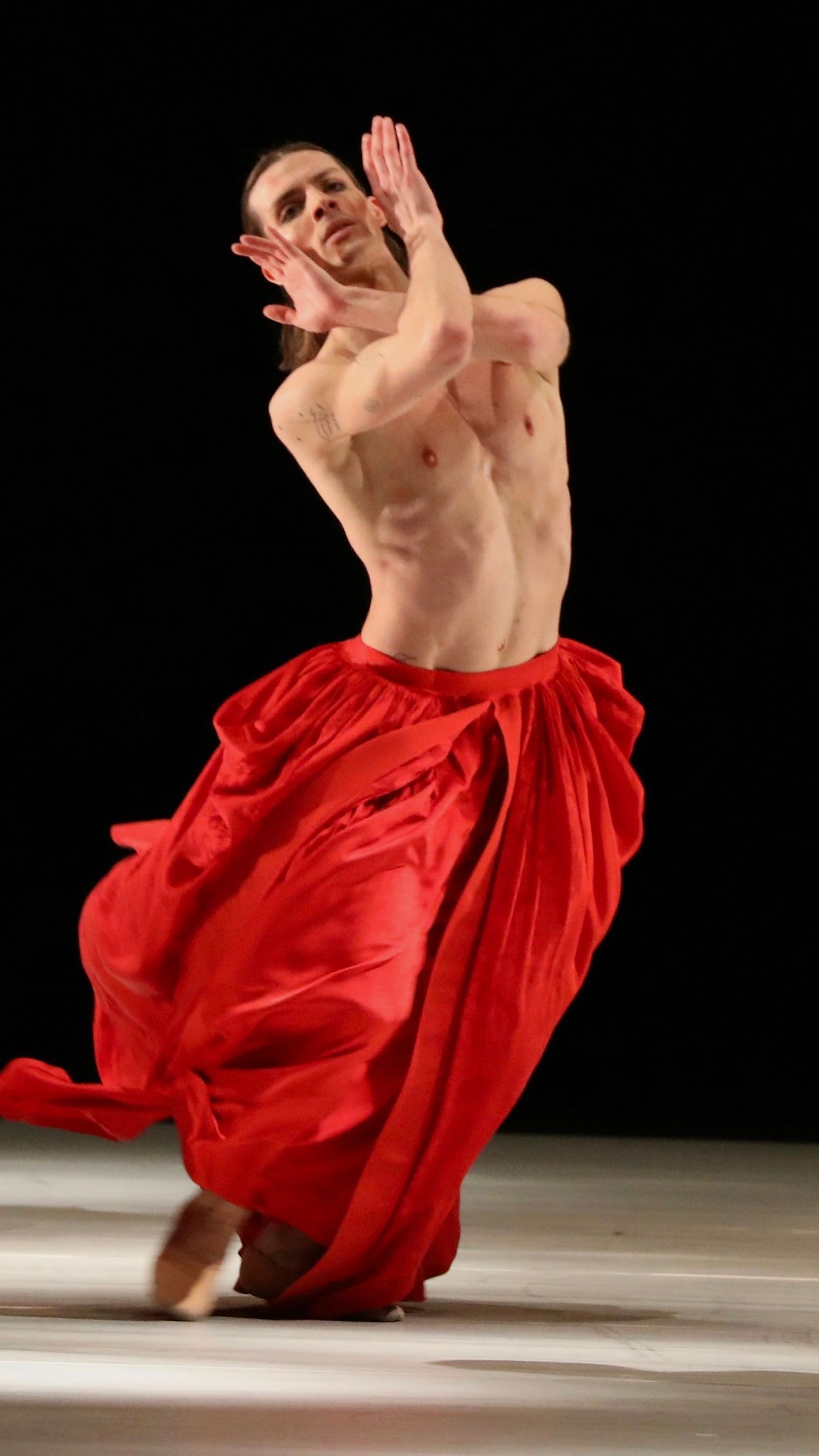 Ballet dancer in red skirt with arms raised, performing on stage with dark background.