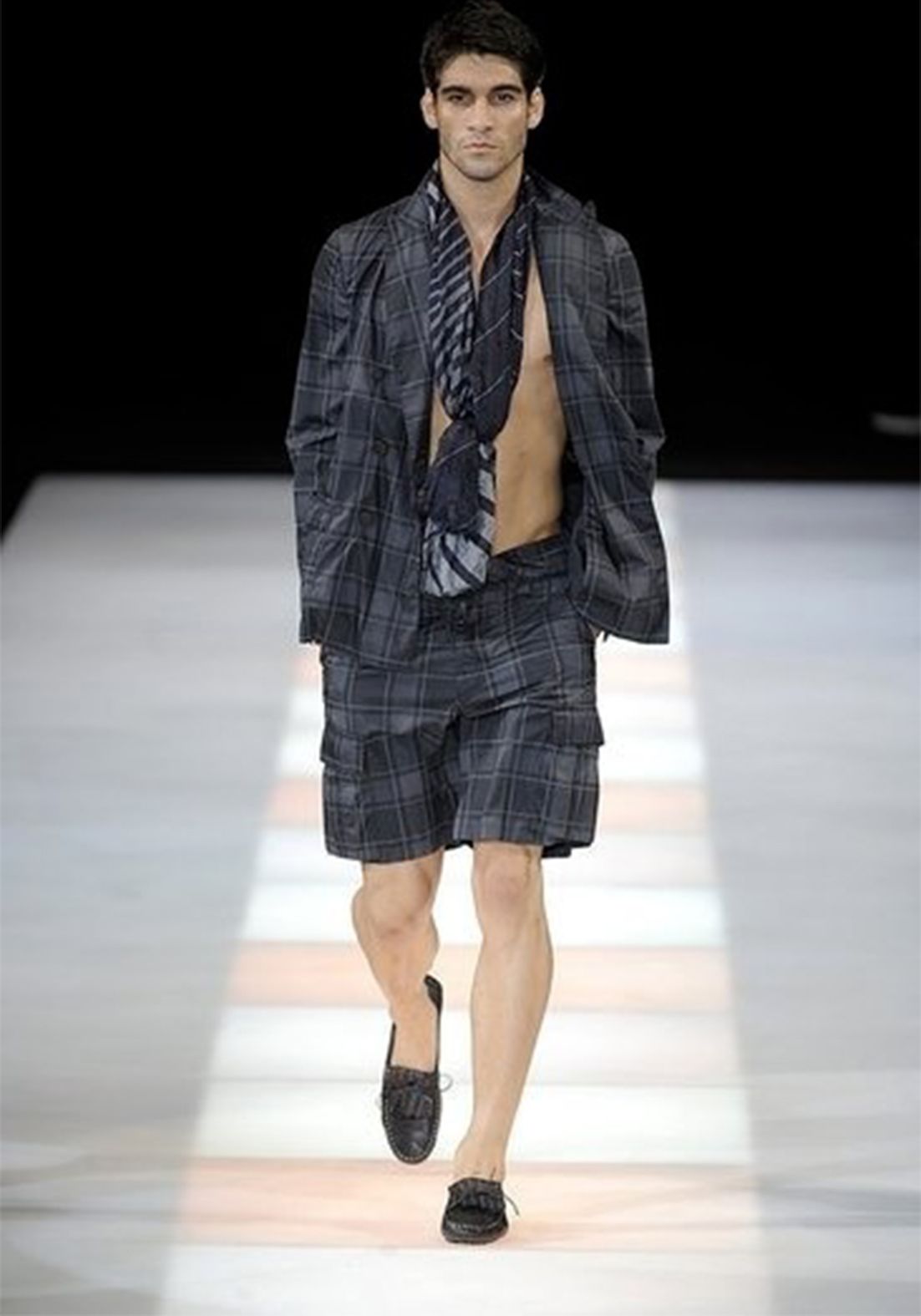 Man in patterned suit jacket, shorts, and scarf, walking a runway with black loafers, bare-chested.