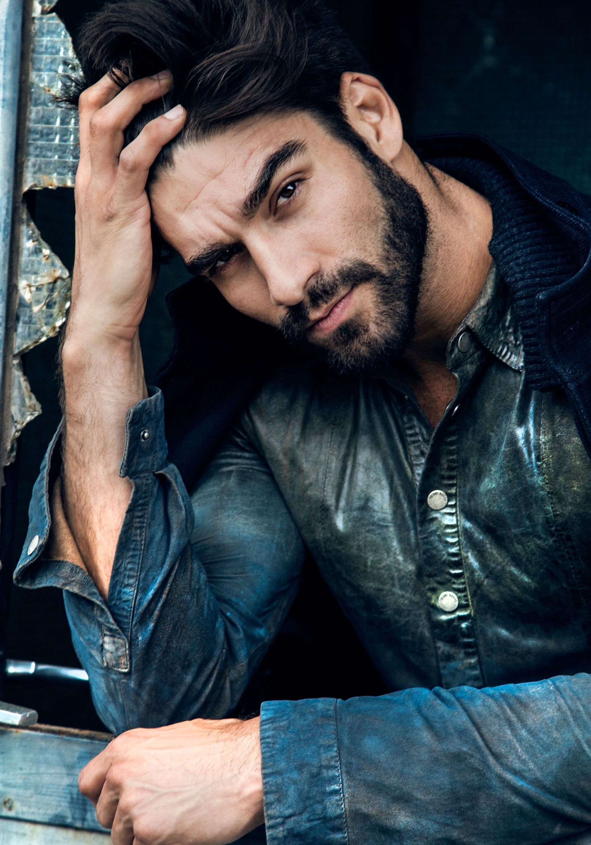Man with dark hair and beard, resting hand on head, looking towards camera. Wearing blue denim shirt.