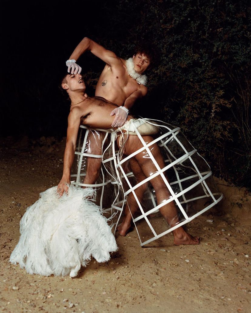 Two shirtless people pose outdoors, wearing cage-like skirts. One reaches toward the other's mouth. Dark, neutral setting.
