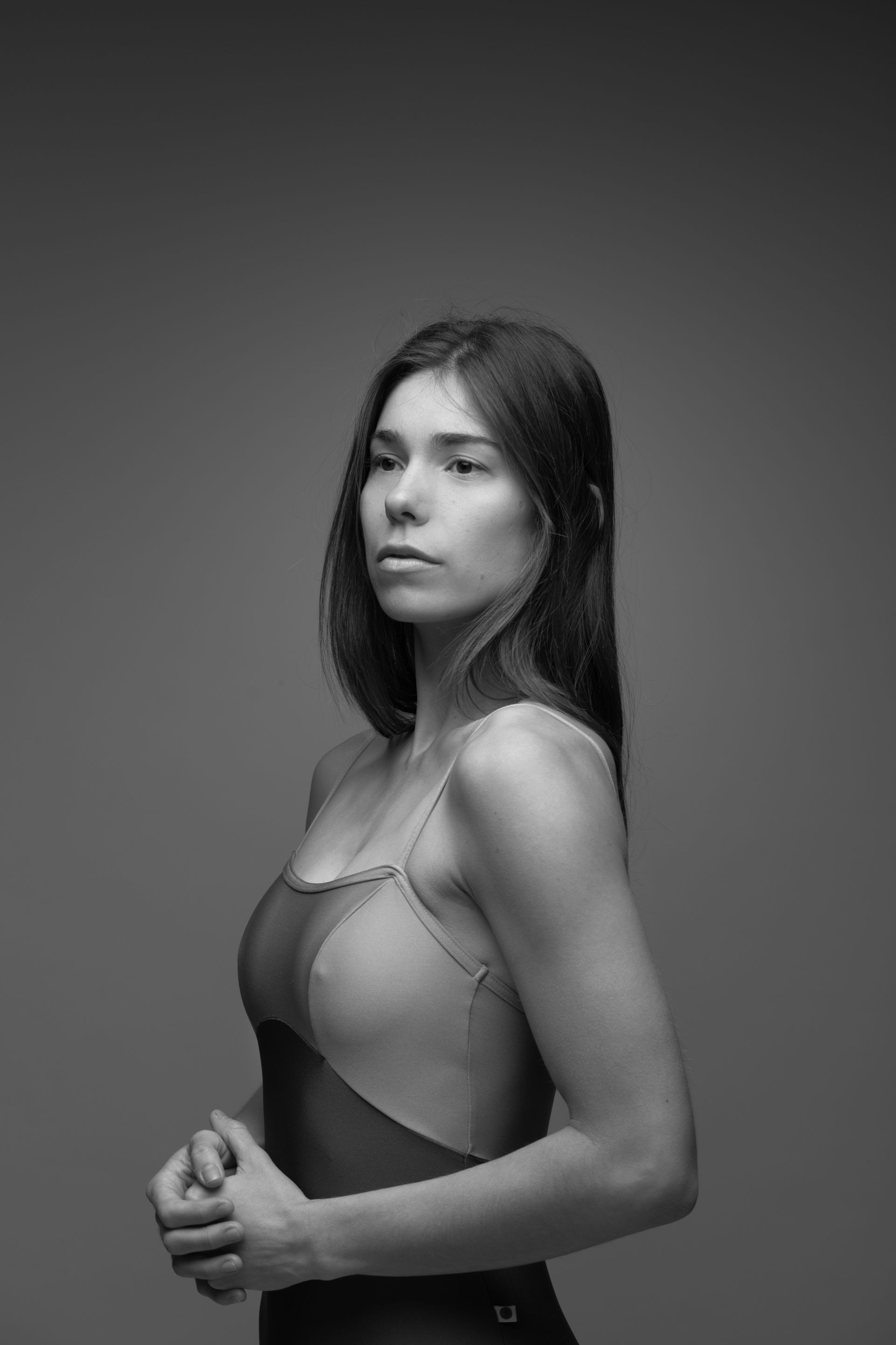 Woman in a leotard, looking away, arms clasped. Gray background, studio setting.