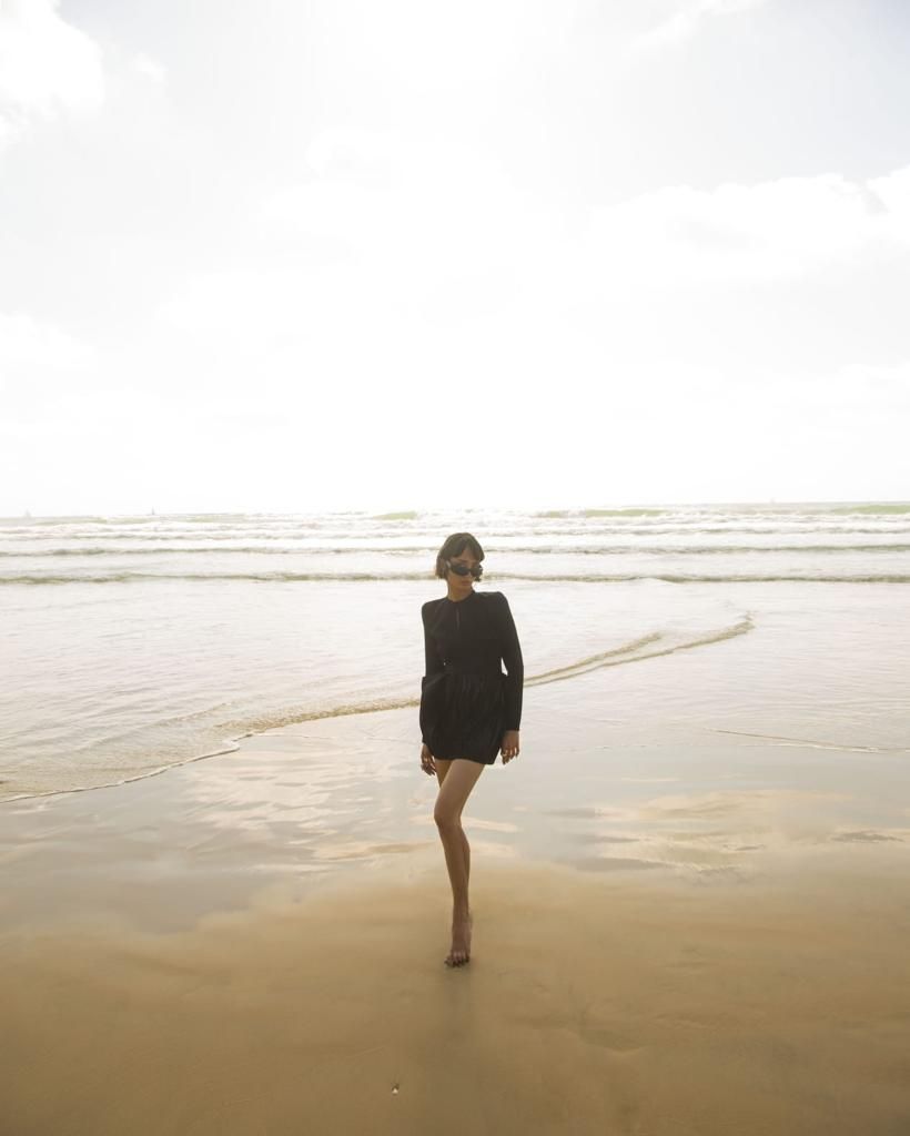 Person in black dress and sunglasses walking on a sandy beach, waves in the background under a bright sky.