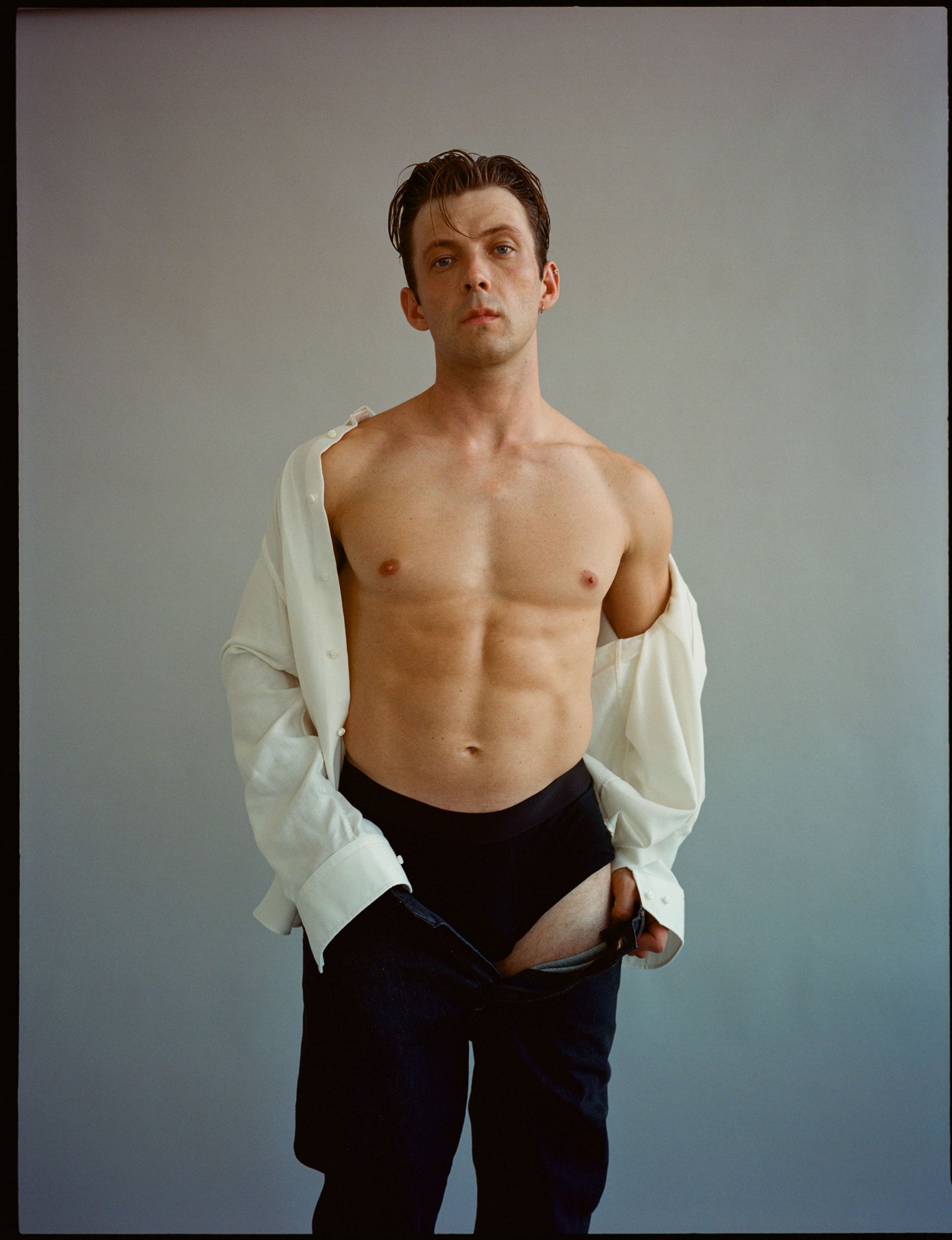 Man with open white shirt, black briefs and pants, torso exposed. Stands against grey background.