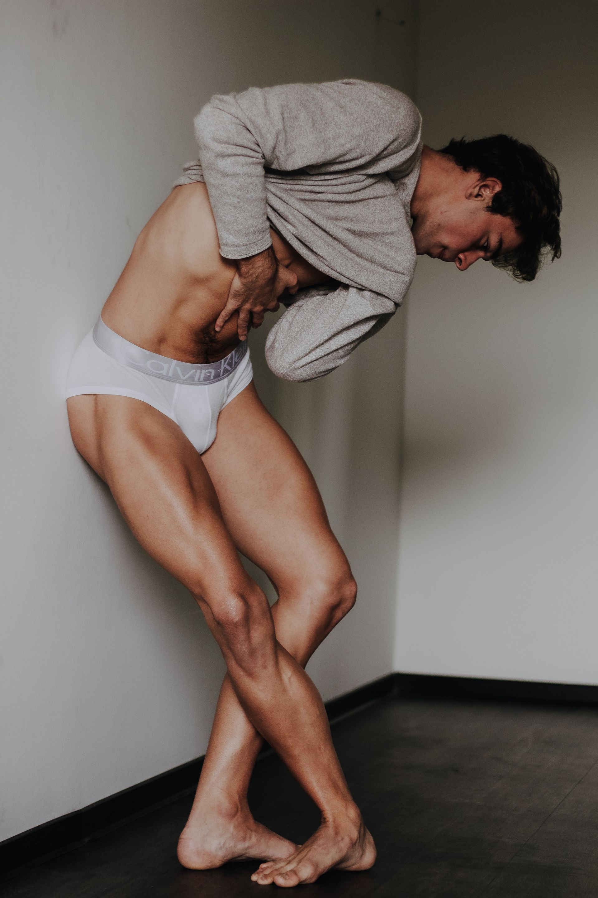 Man leaning against a wall, lifting shirt to reveal abs. Legs crossed, wearing white underwear.