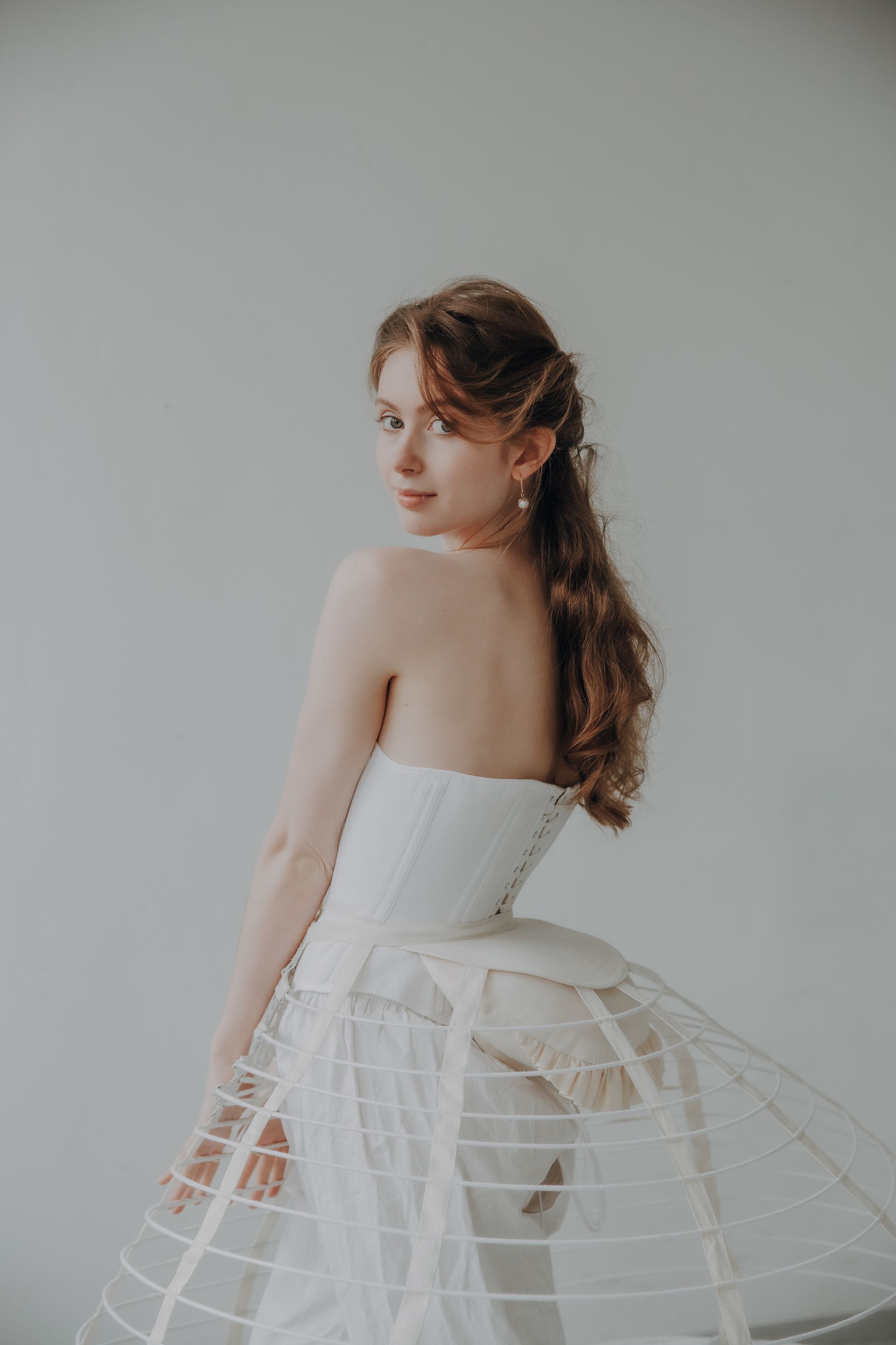 Woman in white corset and hoop skirt, looking over her shoulder against a plain backdrop.