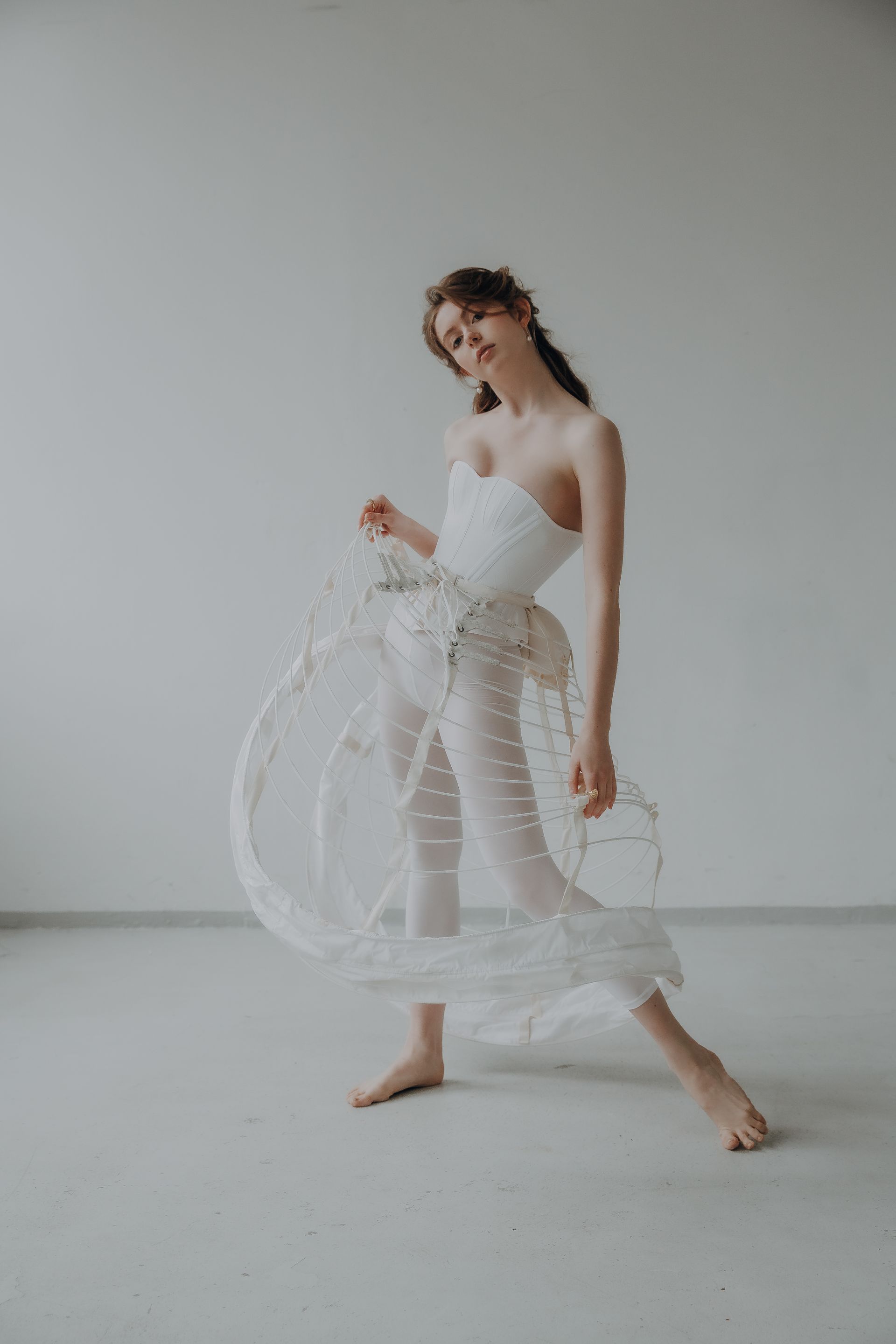 Woman in a white corset and skirt frame stands barefoot, smiling, against a plain white background.