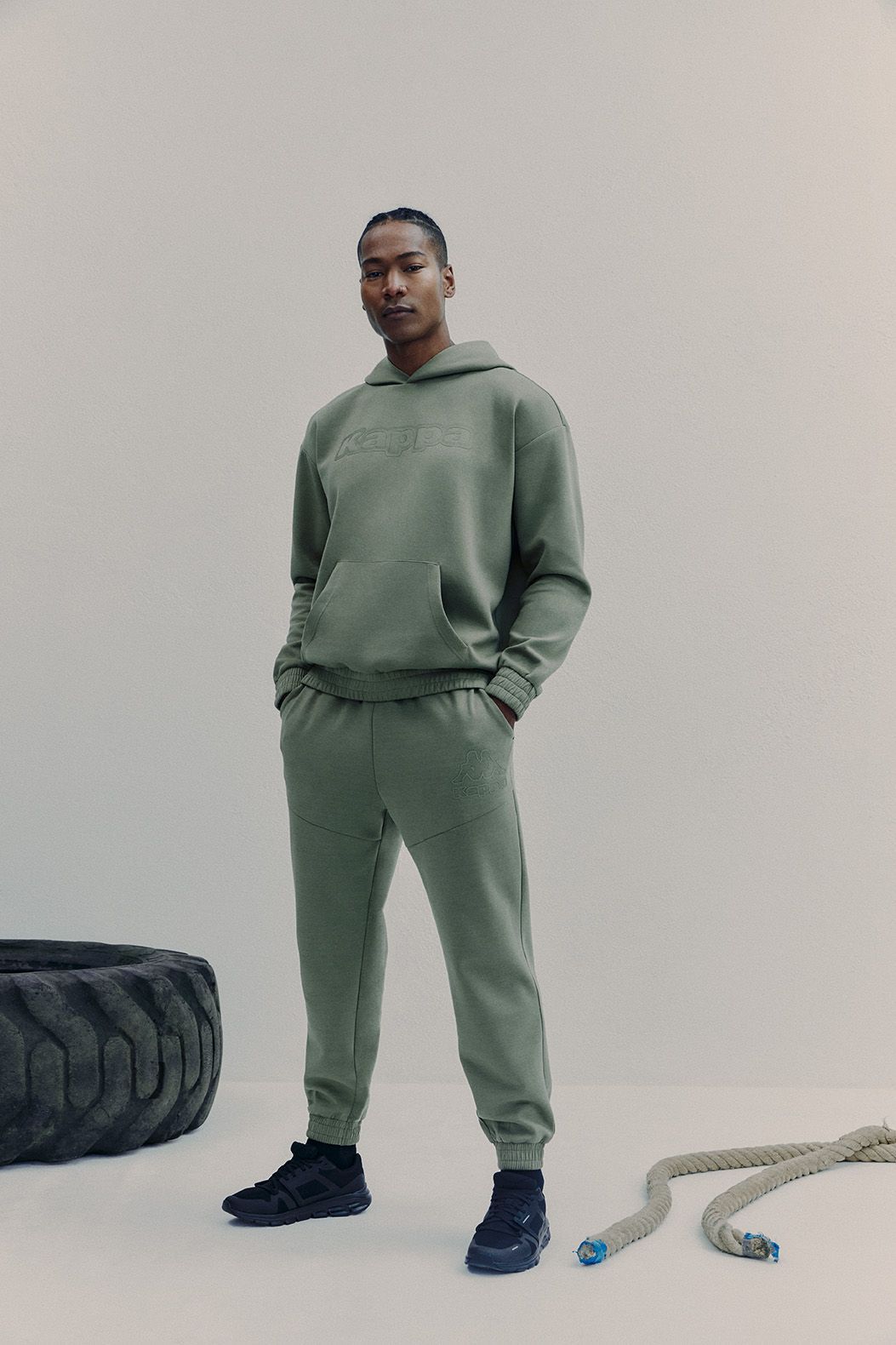 Man in green sweatsuit and black shoes stands near tire and rope.