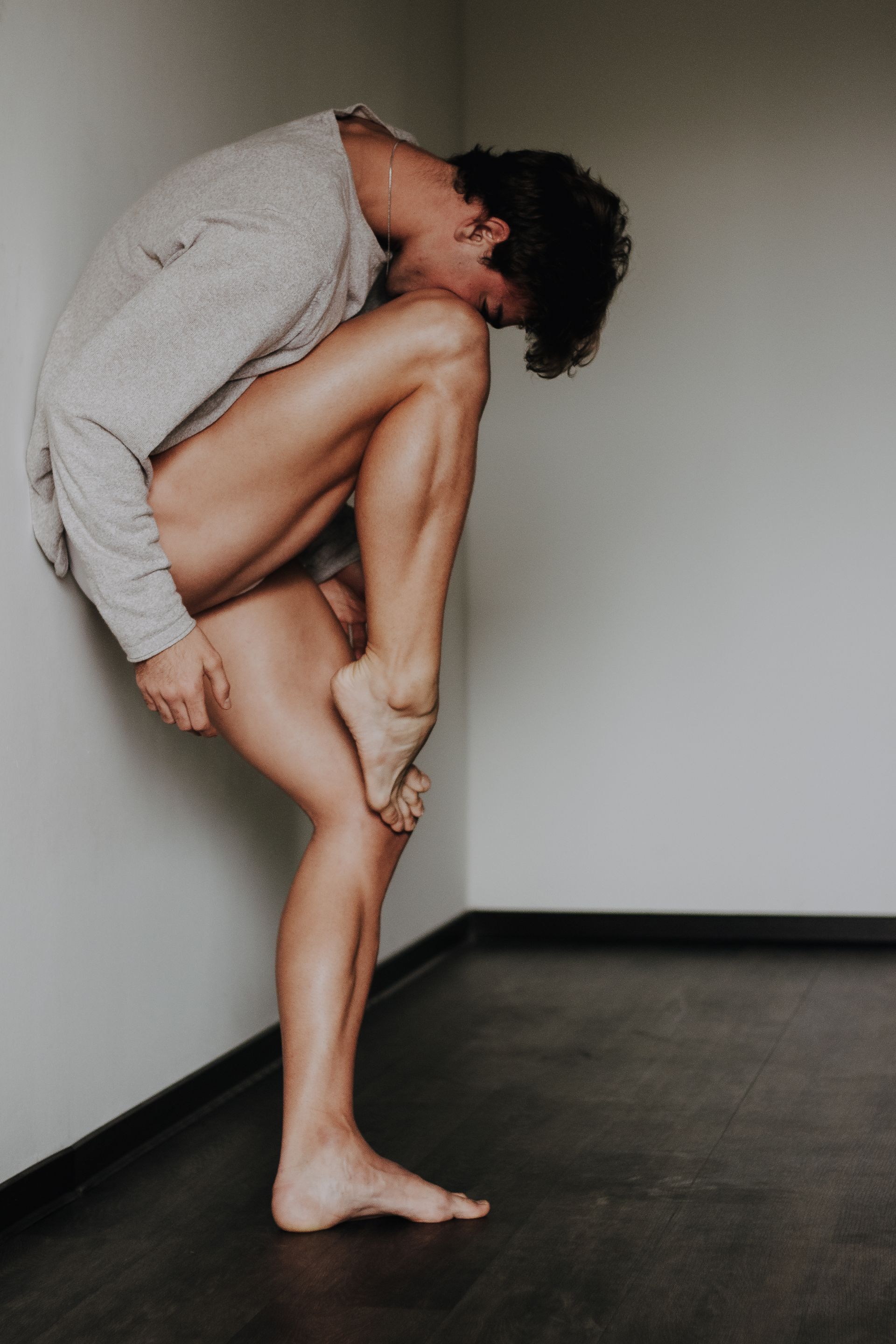 Man in a contorted pose, leaning against a corner wall. He is holding his leg with his hand.