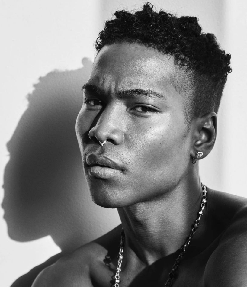 Man with curly hair, nose ring, and chain necklace; looking toward the camera with shadow on the wall.