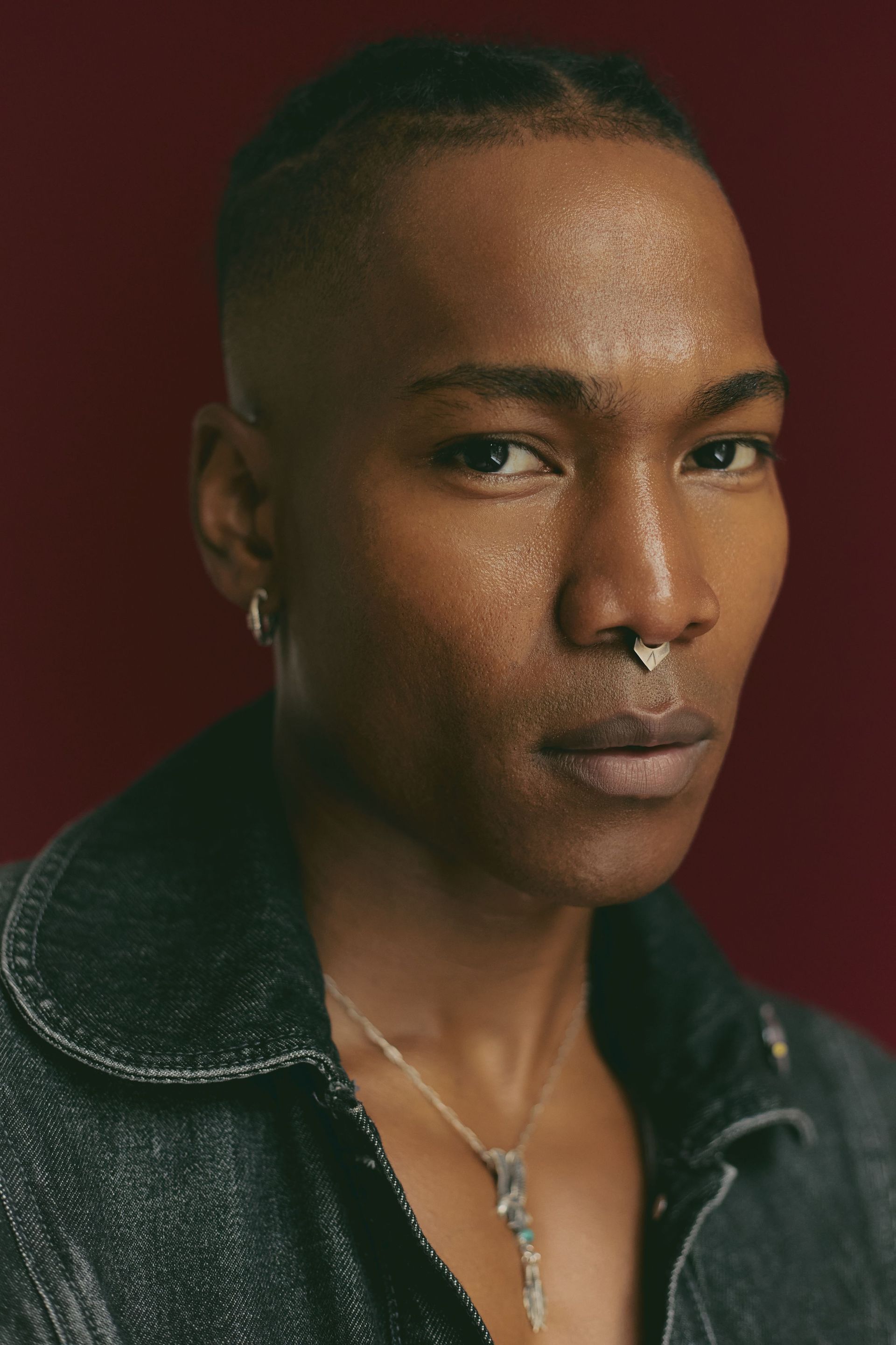 Person with short hair, nose ring, denim jacket, looking at the camera against a burgundy background.