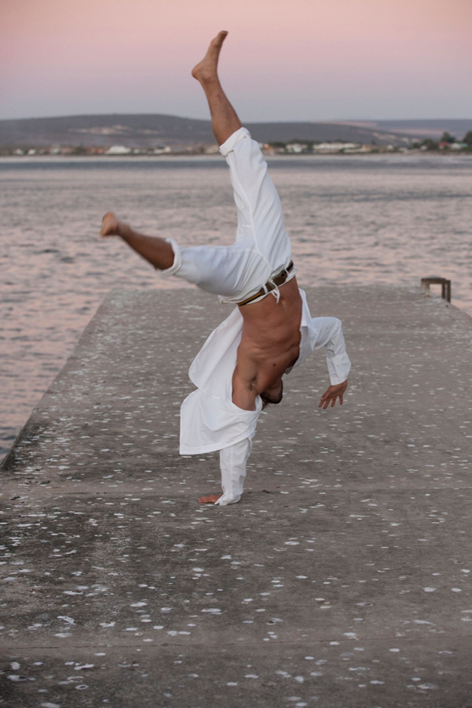 Man doing a handstand on a pier near water; wearing white clothing, sunset in the background.