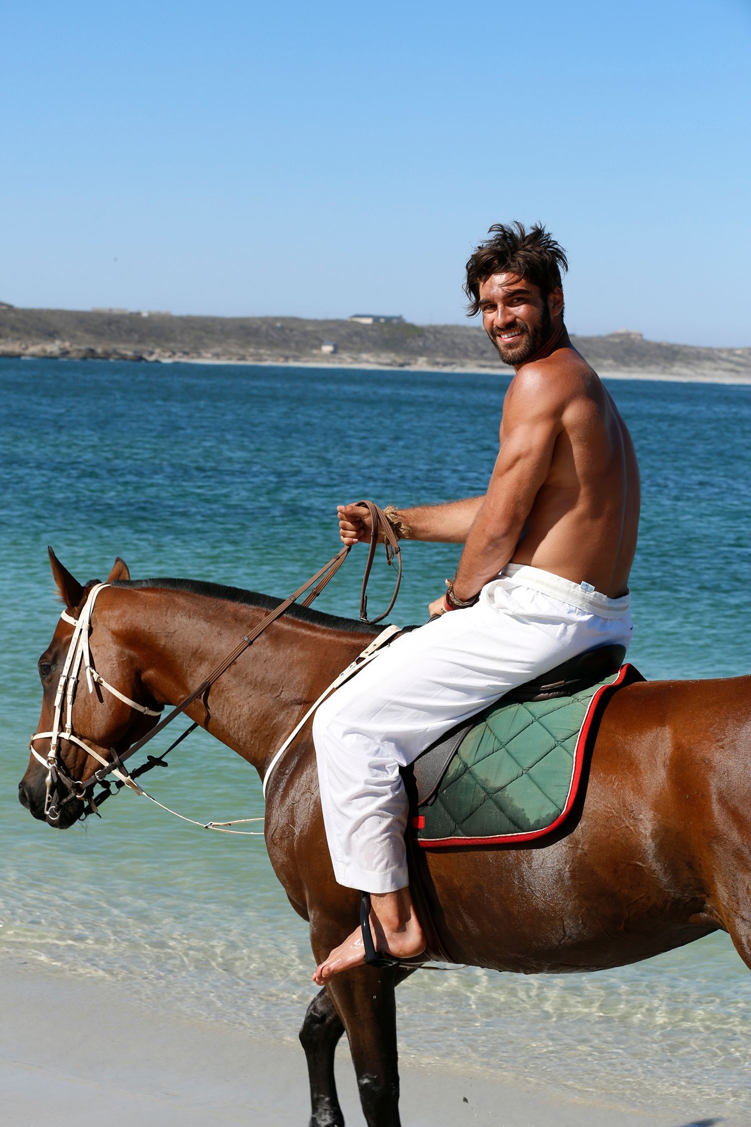 Man rides a brown horse in shallow ocean water. He smiles, shirtless, wearing white pants.