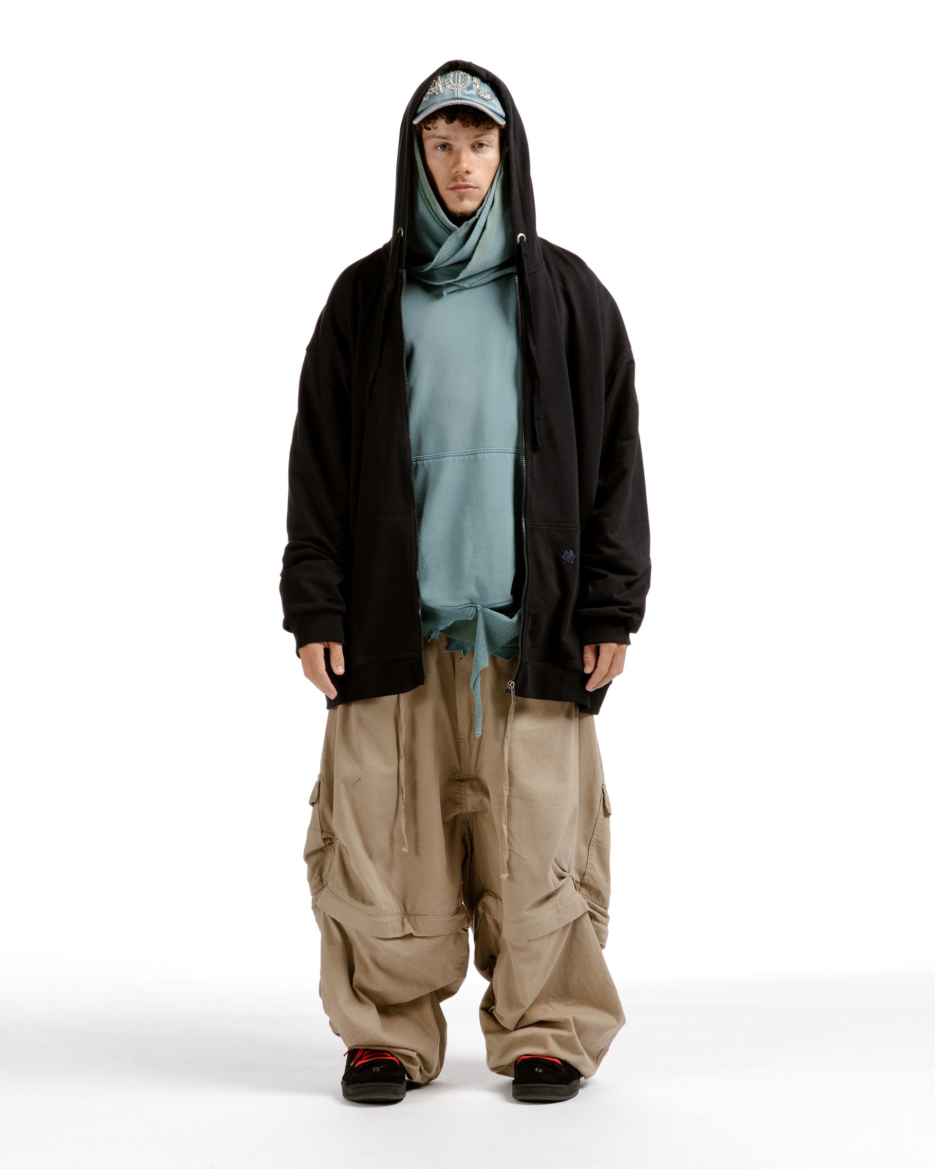 Person wearing a black hooded jacket, blue hoodie, tan cargo pants, and a patterned cap.