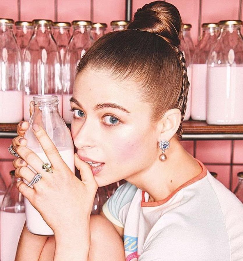 Woman with shaved head, holding a milk bottle, biting her lip, with a rack of milk bottles in the background.
