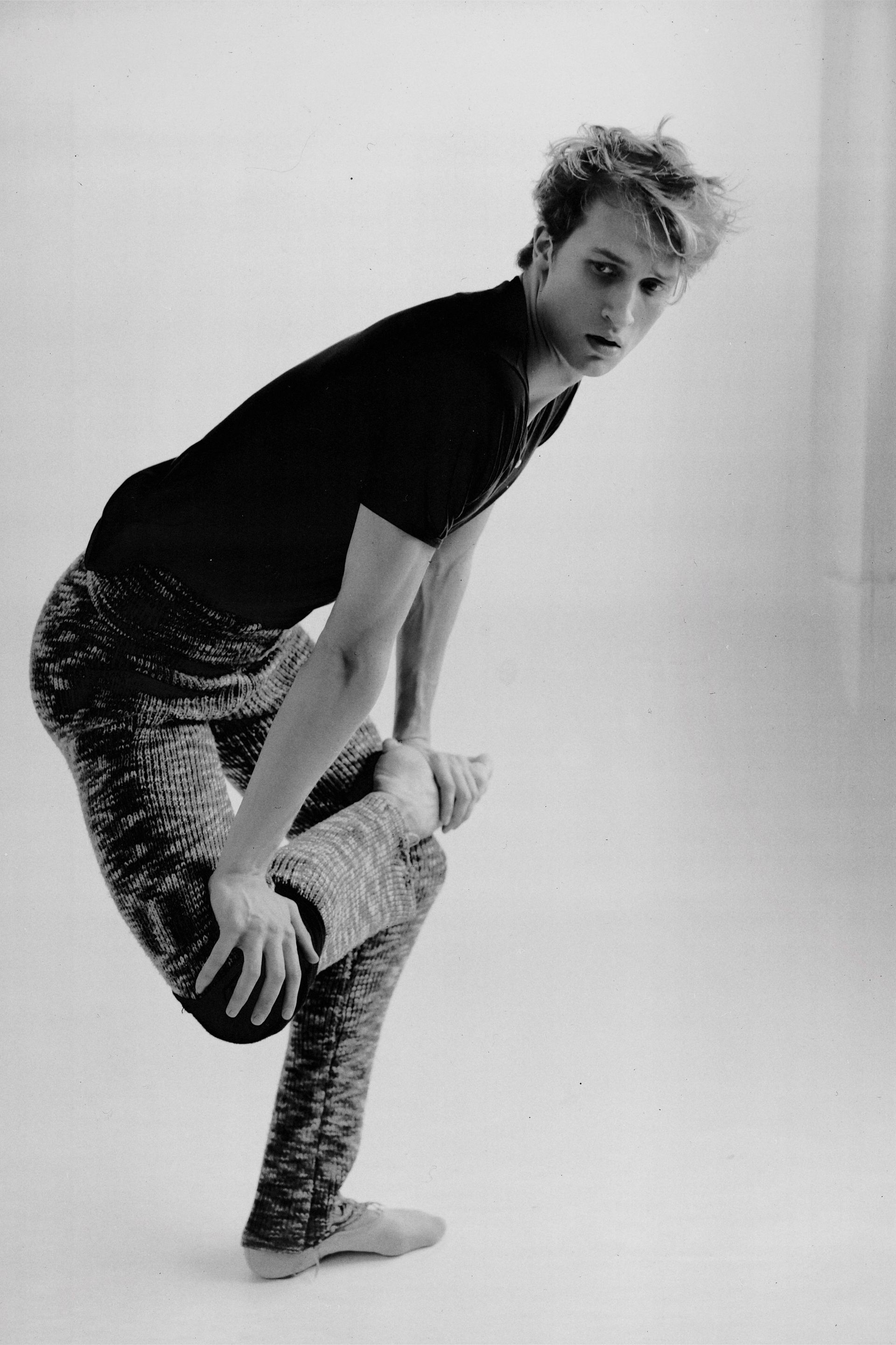 Man in athletic pose, wearing dark shirt and patterned pants, in a studio with neutral background.