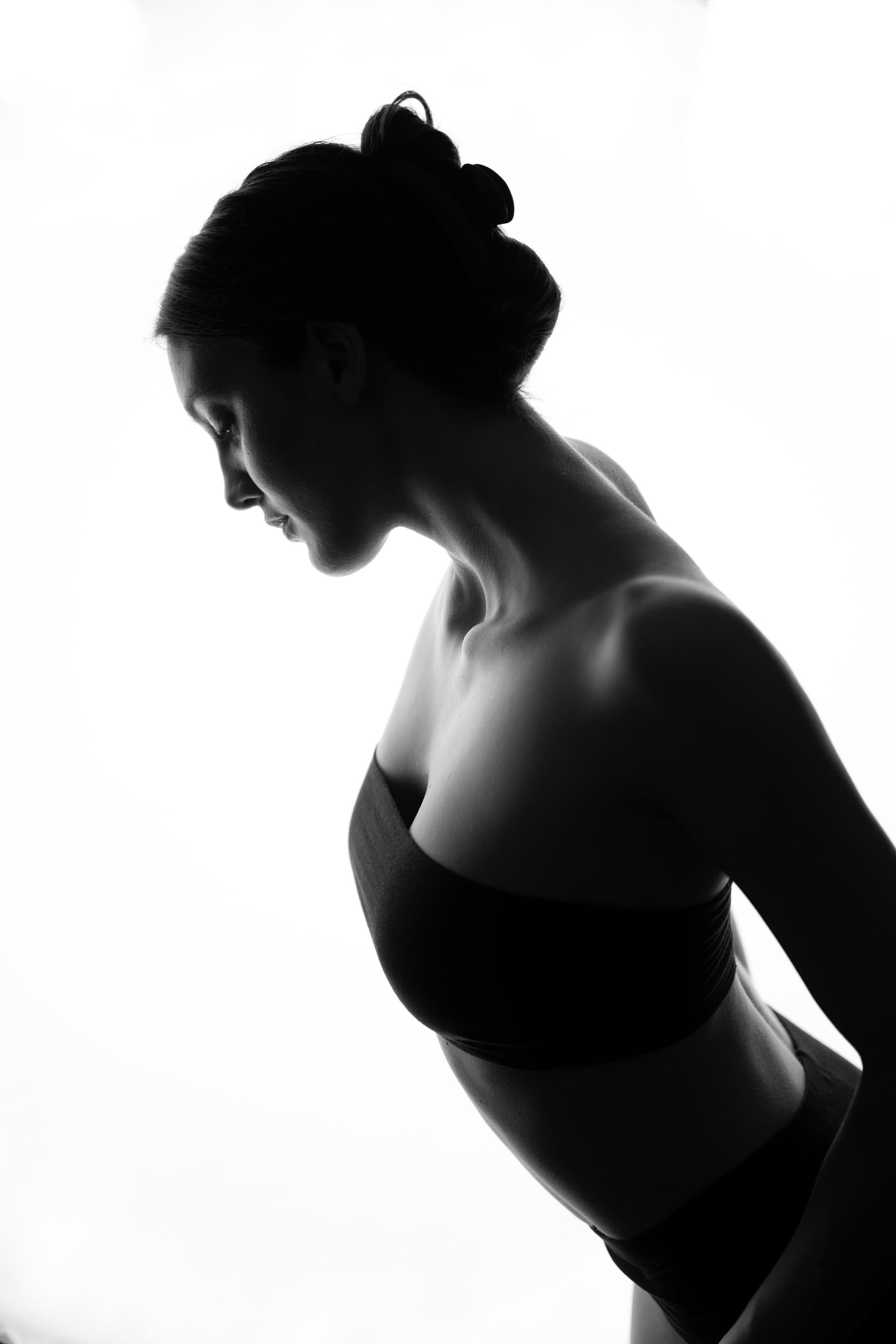 Silhouette of a woman in a strapless top, head tilted down, hair up, against bright background.