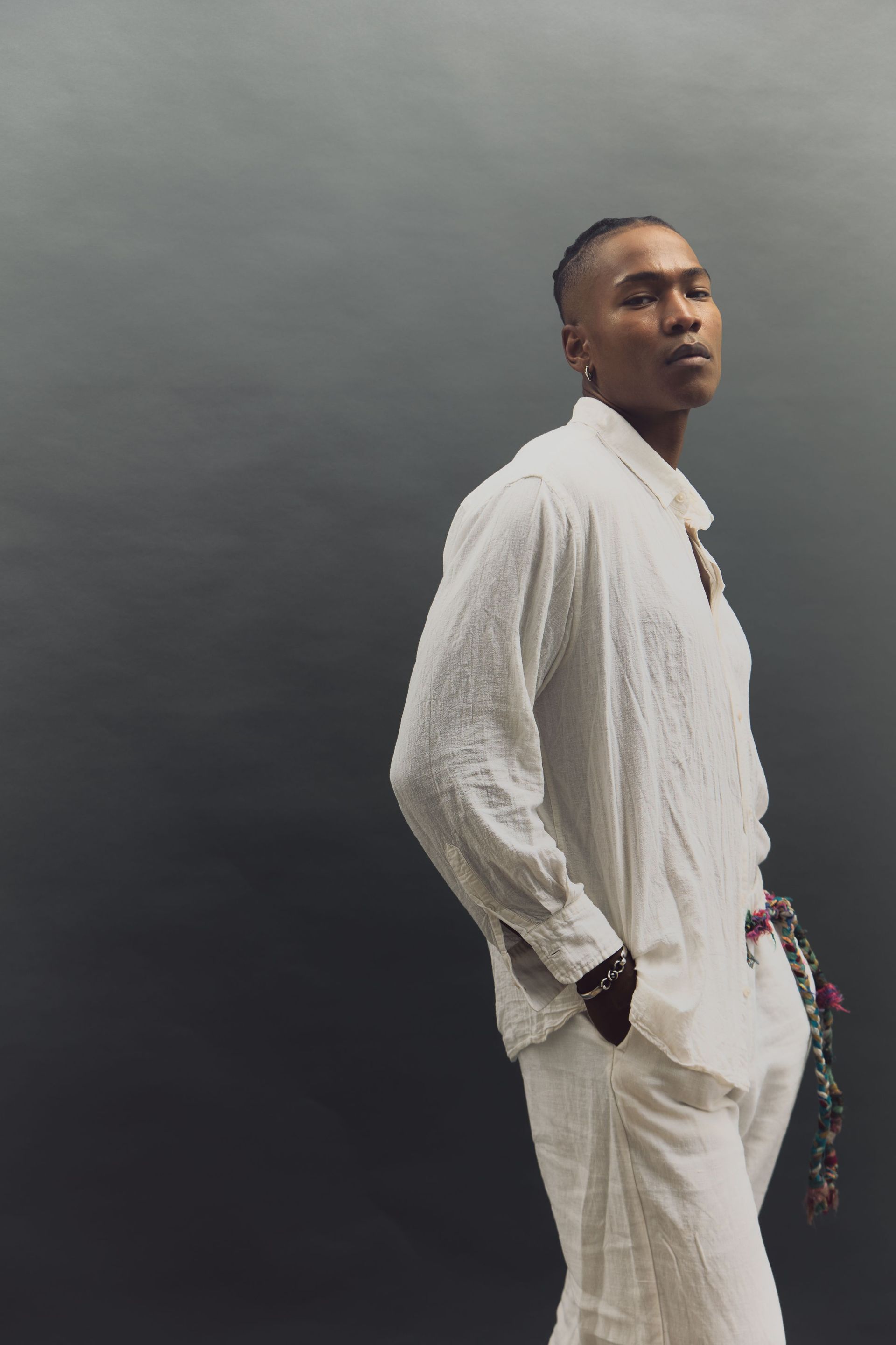 Man in white linen outfit, looking upwards, standing against a gray background.