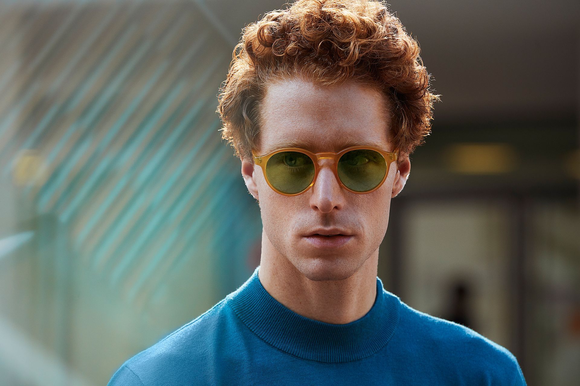 Man with curly red hair wearing yellow-framed sunglasses, blue shirt, outdoors.