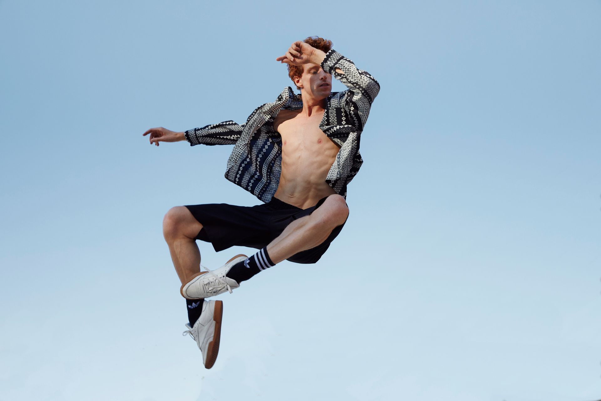 Man in mid-air, squinting, wearing a patterned jacket, shorts, and sneakers against a blue sky.