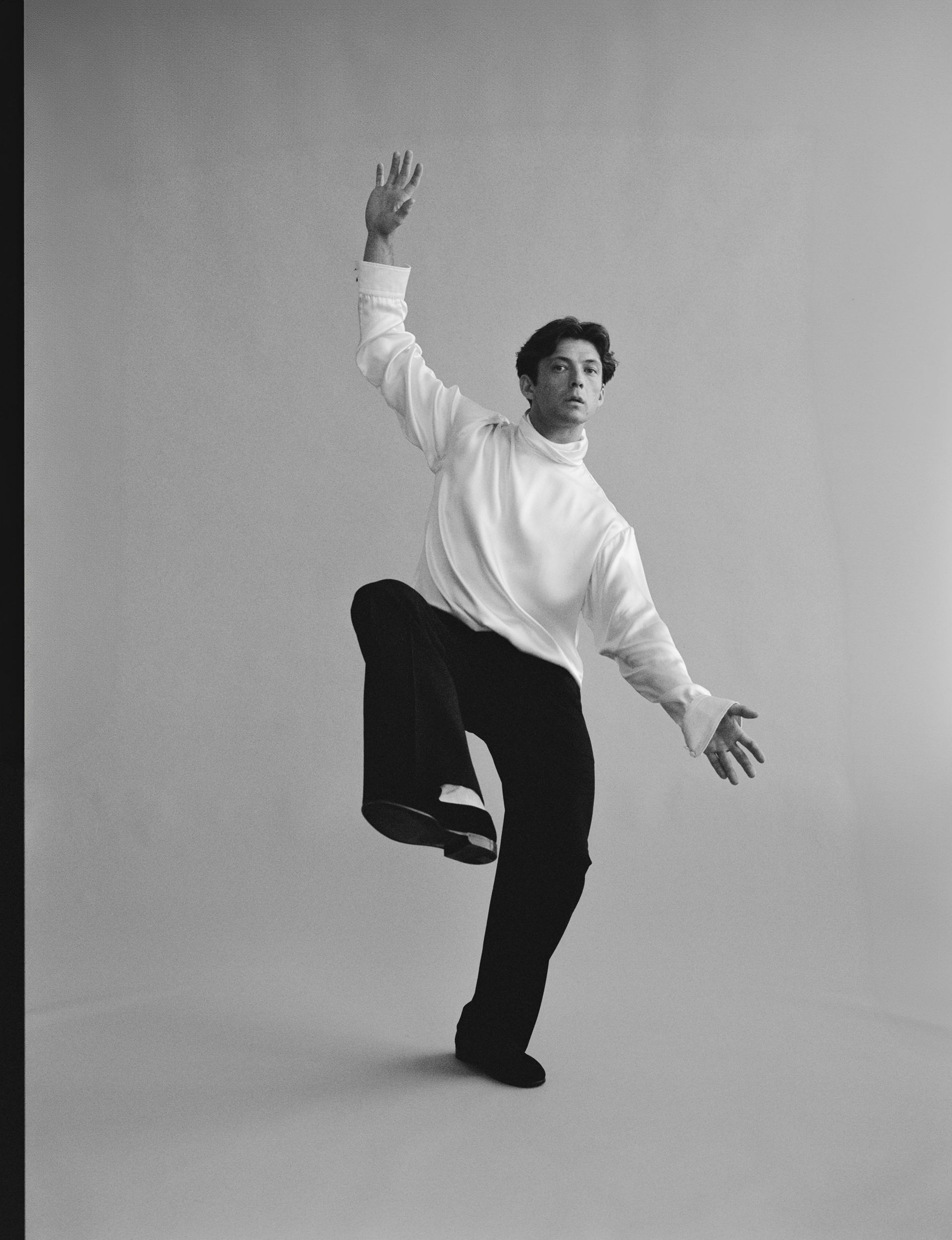 Person in a white turtleneck and black pants balancing in a dance pose against a light gray backdrop.