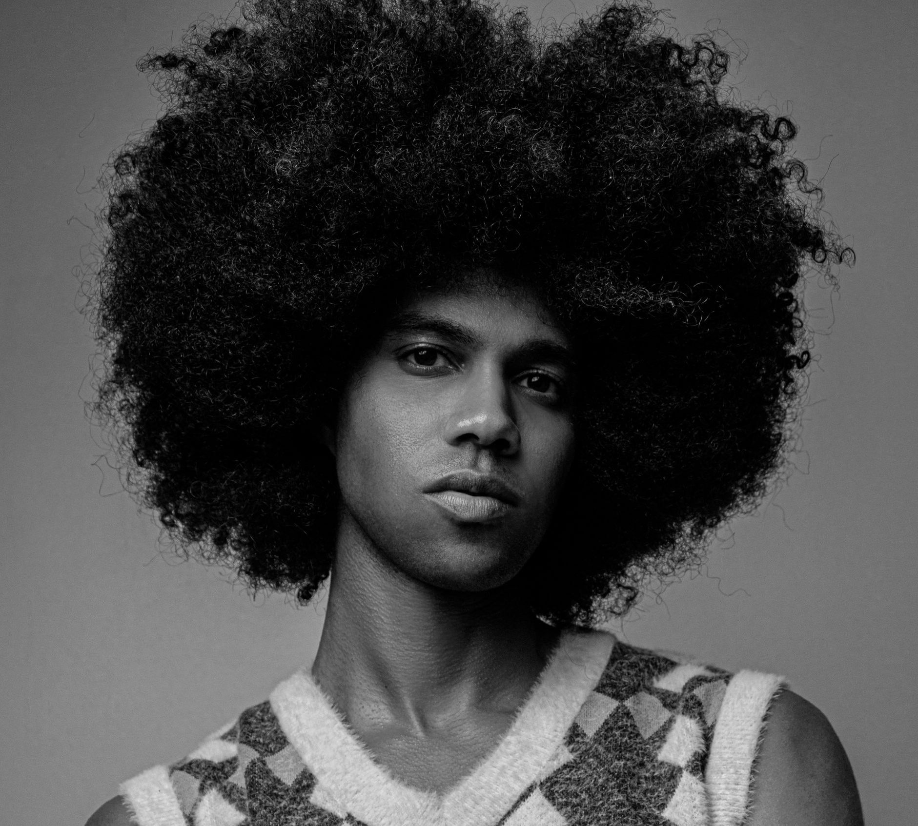 Man with large afro, wearing patterned vest, looking directly at the viewer. Black and white.