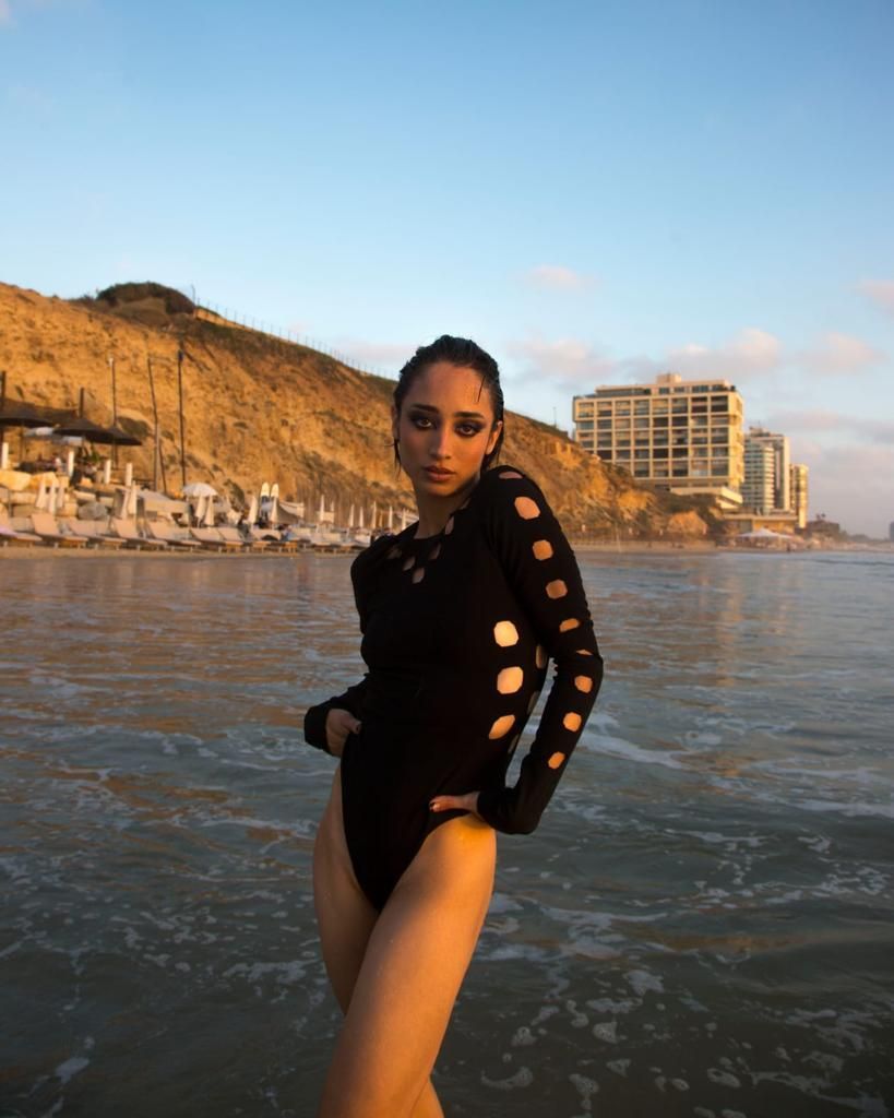 Woman in black bodysuit, standing in shallow water, beach setting with buildings and cliffs.