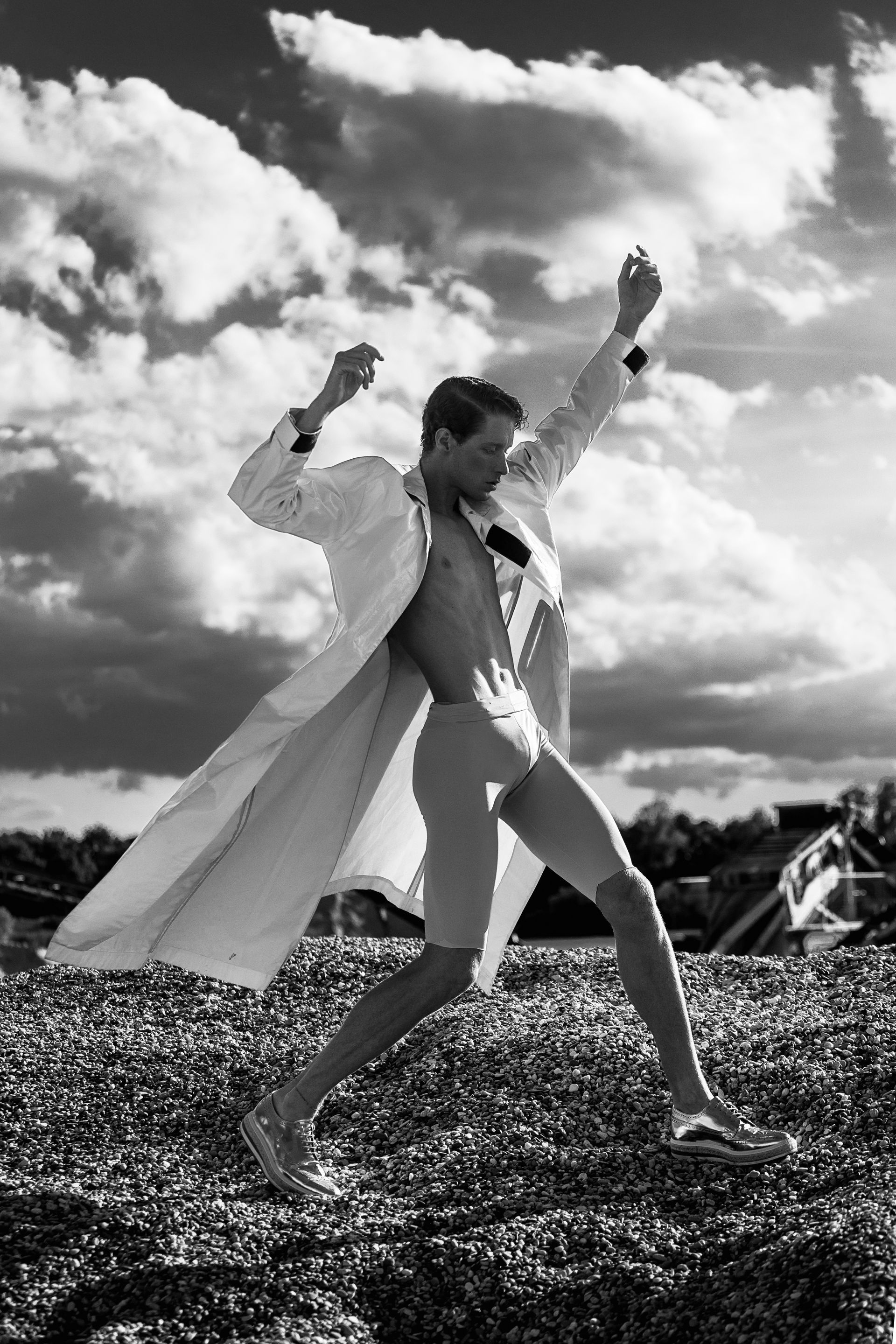 Man in white coat and shorts leaps on a pebbled beach with arms raised against a cloudy sky.