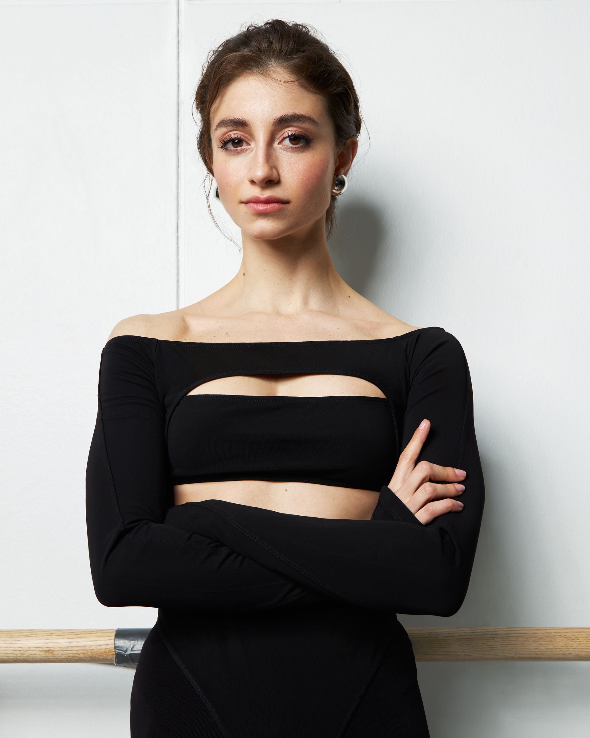 Woman in black cutout top, arms crossed, leaning against a wall with a ballet barre.