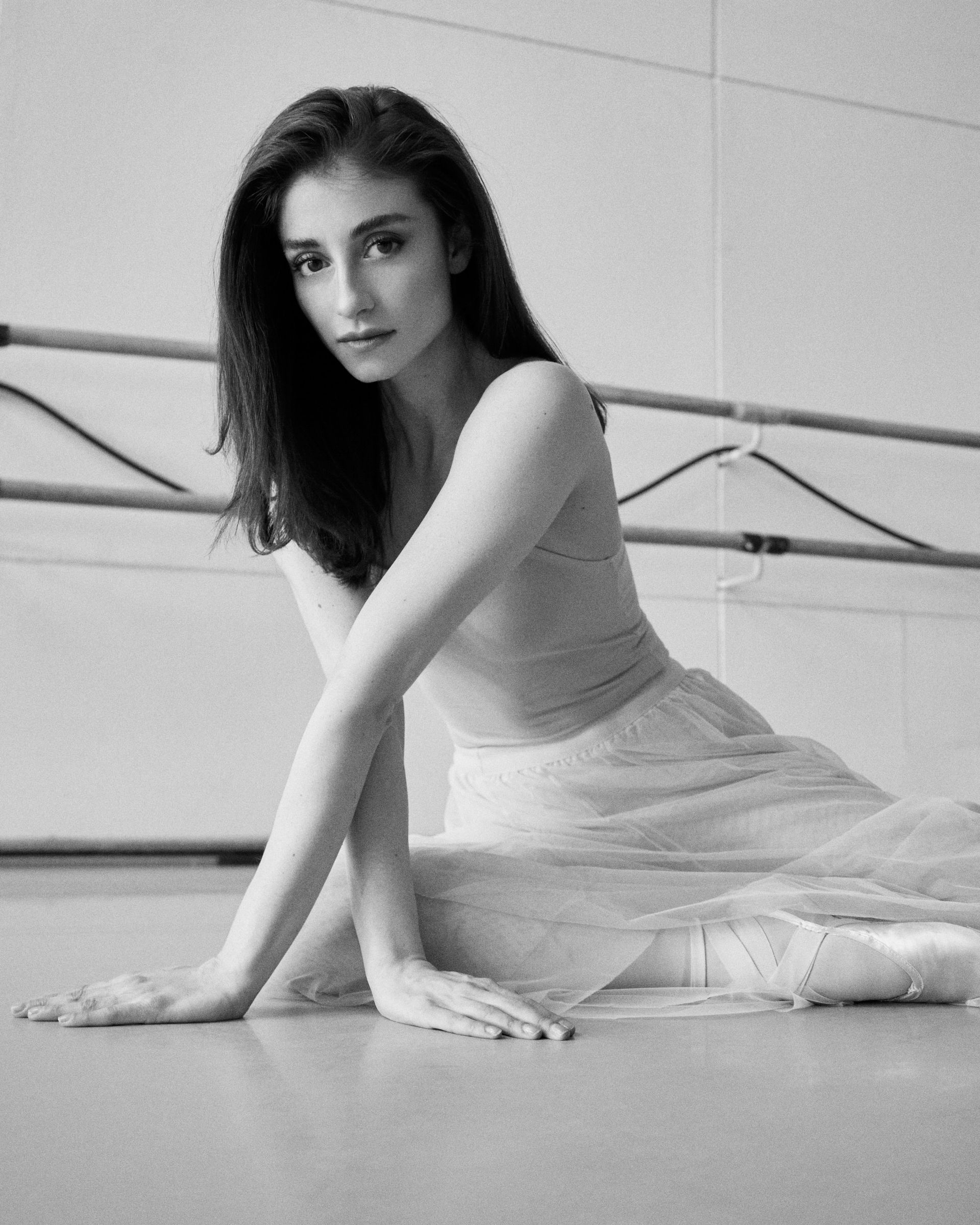 Ballerina sits on floor, arms extended, wearing a tulle skirt in a dance studio.