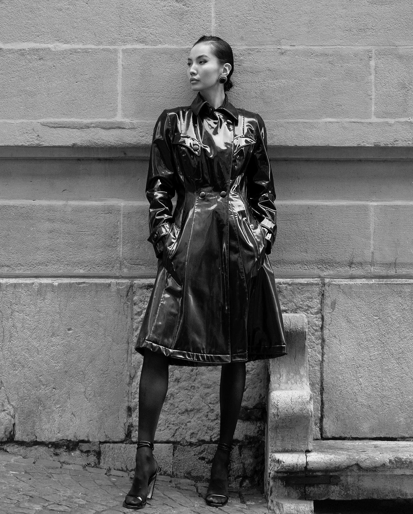 Woman in shiny black coat and ankle boots, leaning against a stone wall, looking away.