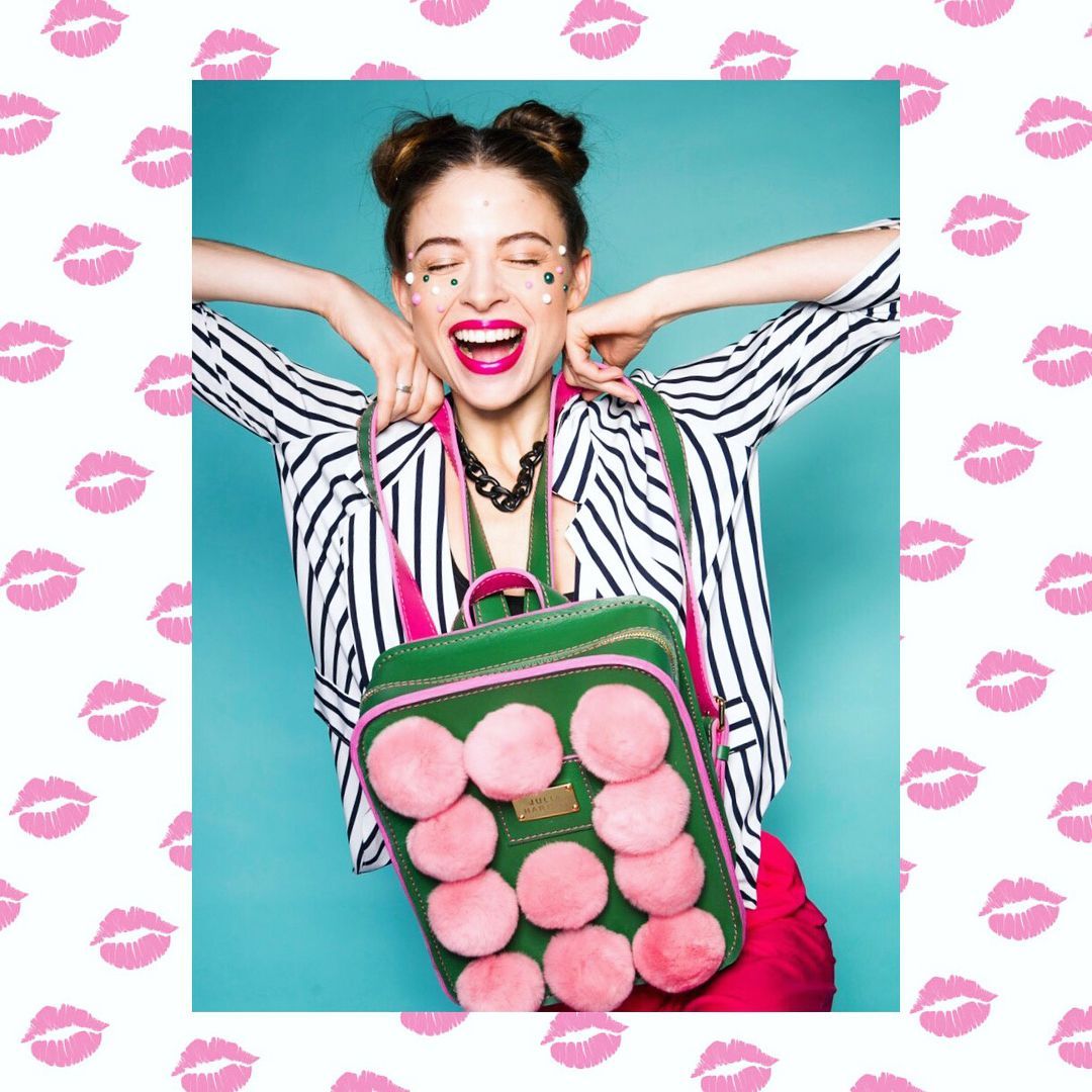 Woman smiling, holding a green backpack with pink pompoms, in a white and black striped jacket.