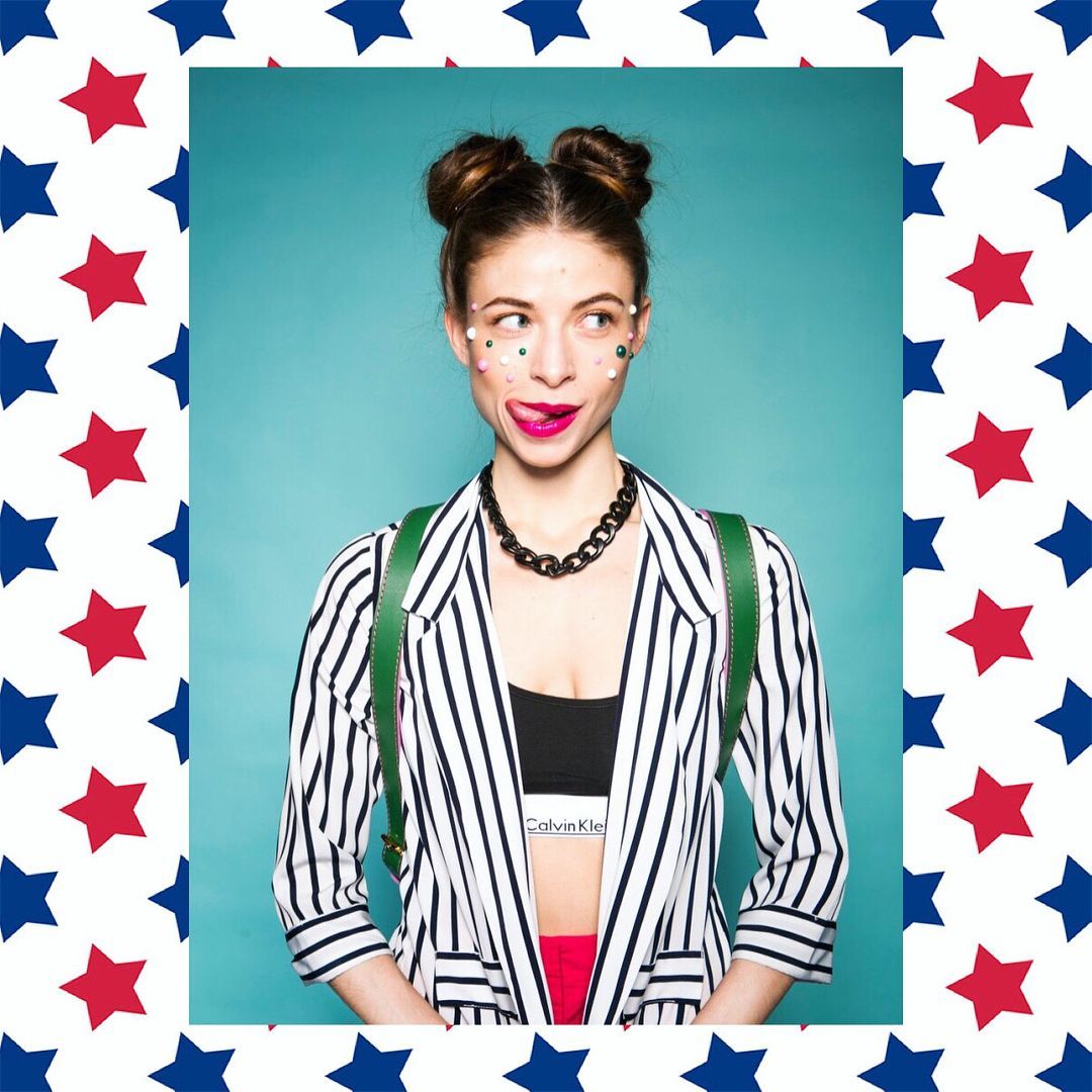 Woman with double buns, striped blazer, and stars on her face sticks out her tongue.