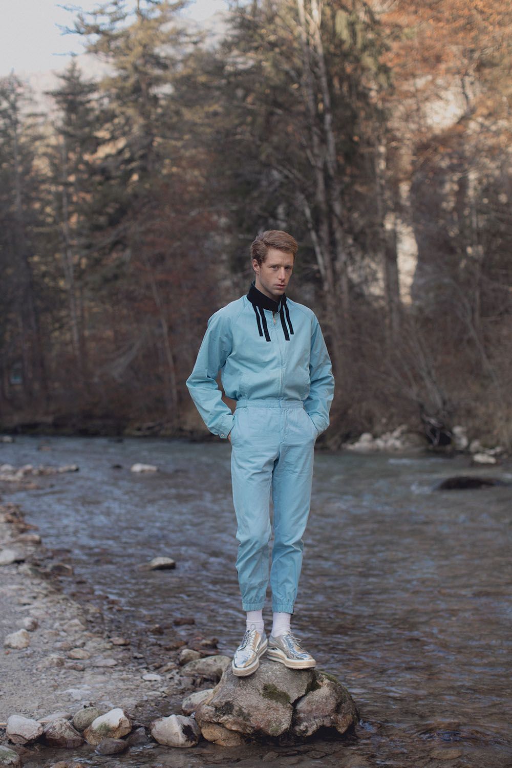 Person in blue jumpsuit stands on a rock by a stream in a forest.