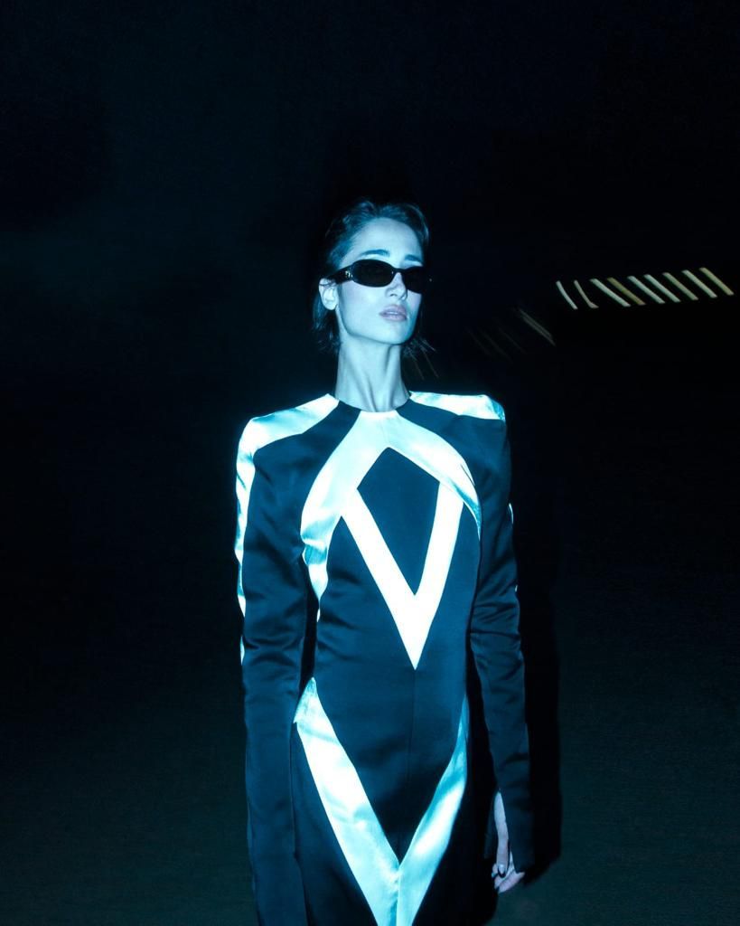 Woman wearing sunglasses and a black jumpsuit with white geometric accents in a dark setting.