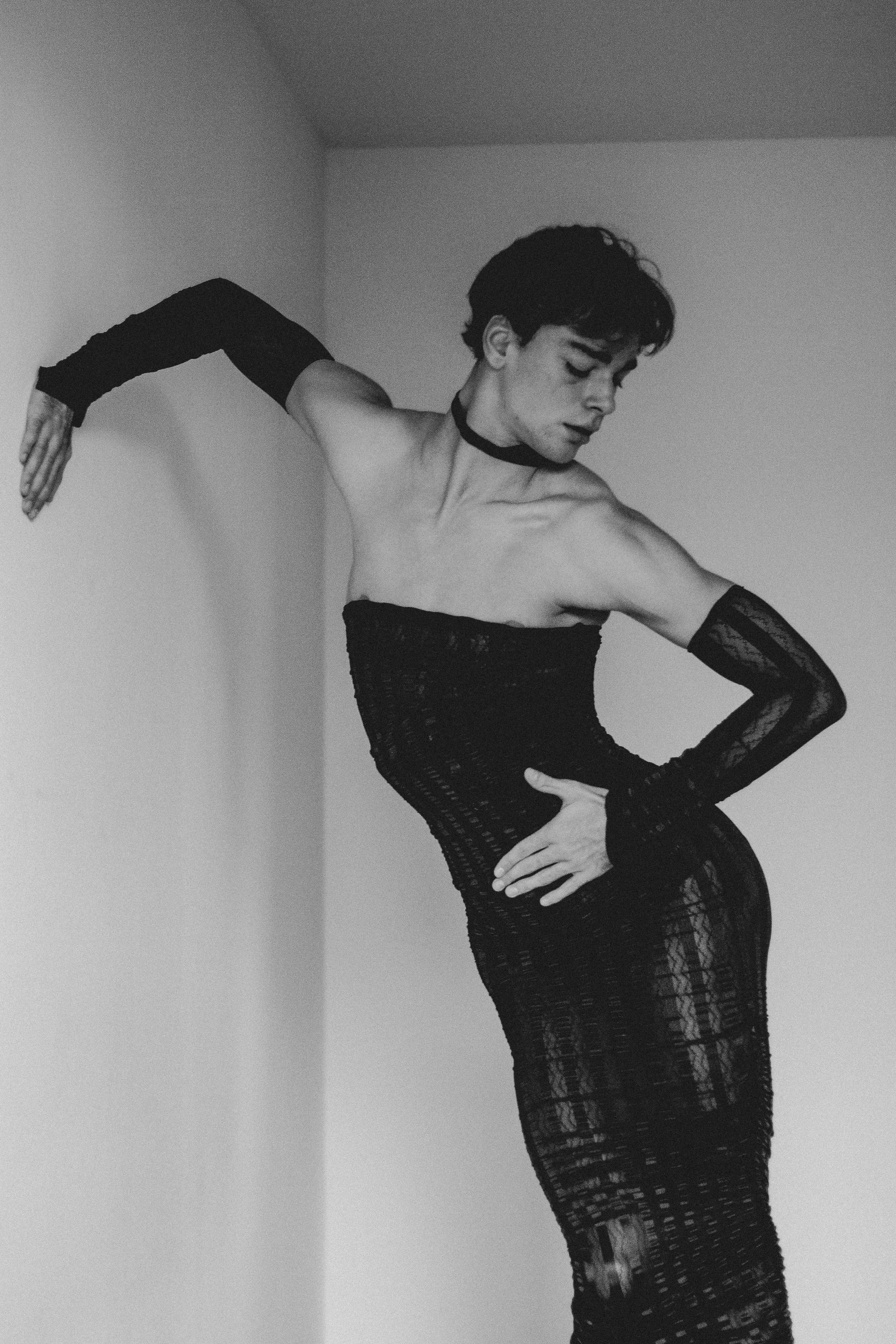 Person in black dress and arm gloves leans against a white wall, hand on waist, looking down.