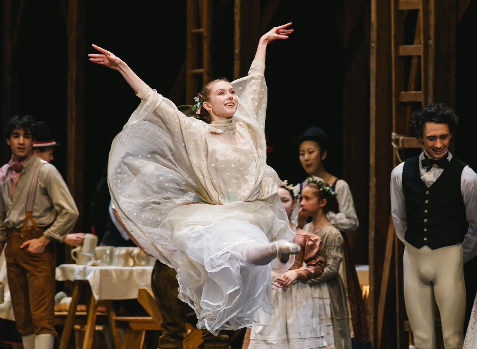 Ballerina in white dress leaping onstage, arms raised, surrounded by other dancers and set.