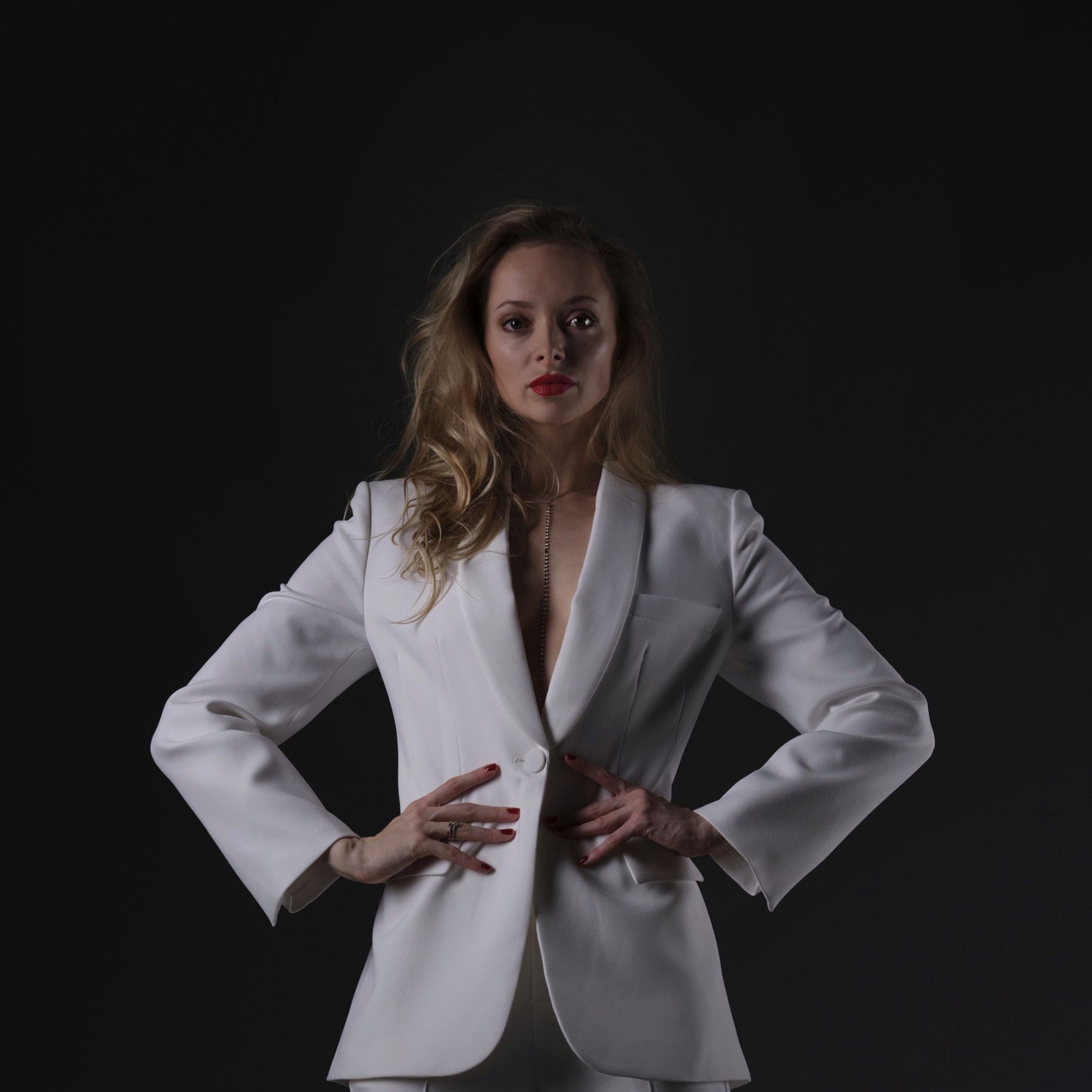 Woman in white suit, hands on hips, looking at camera. Red lipstick. Dark background.