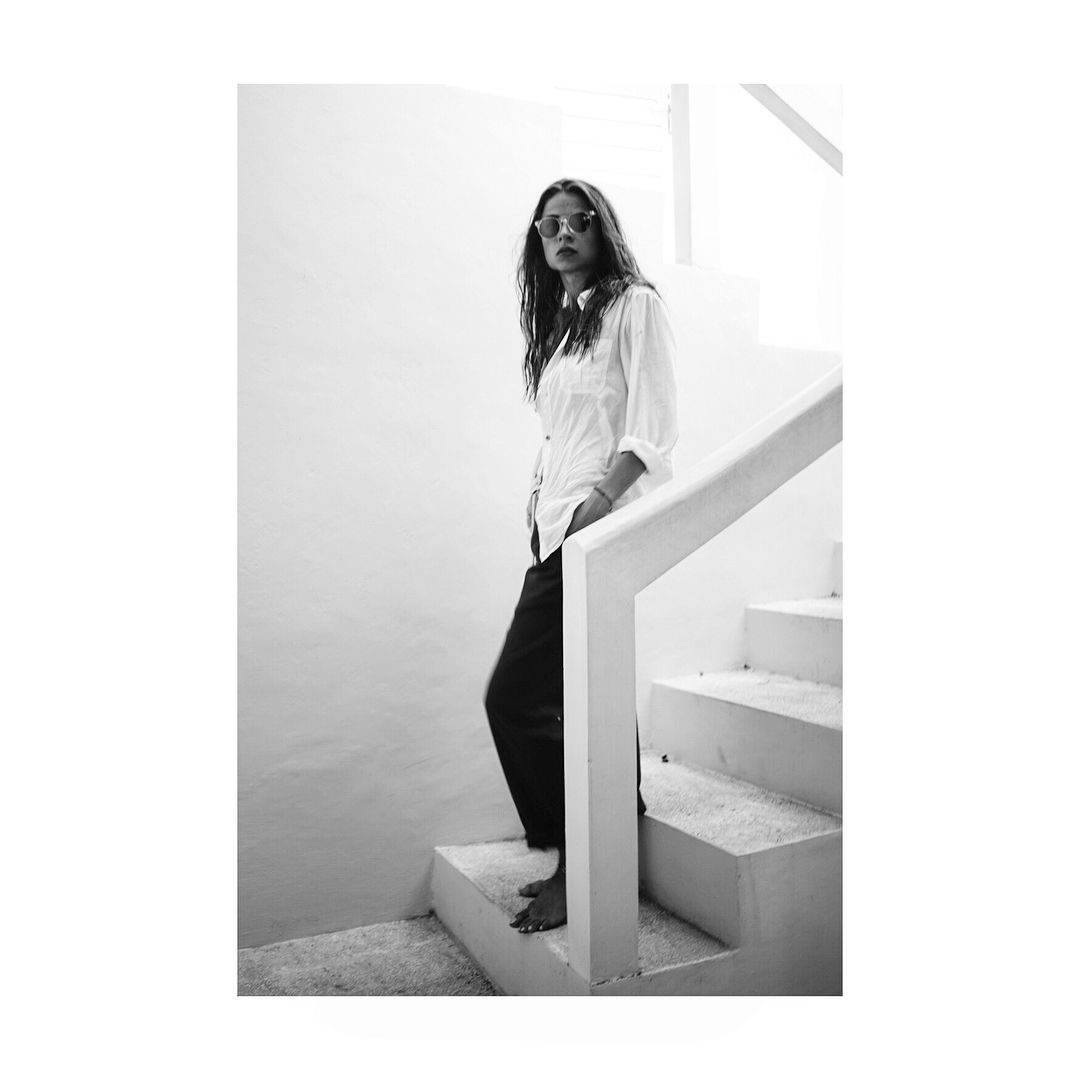 Woman in white shirt and dark pants stands on a white staircase, barefoot with sunglasses.